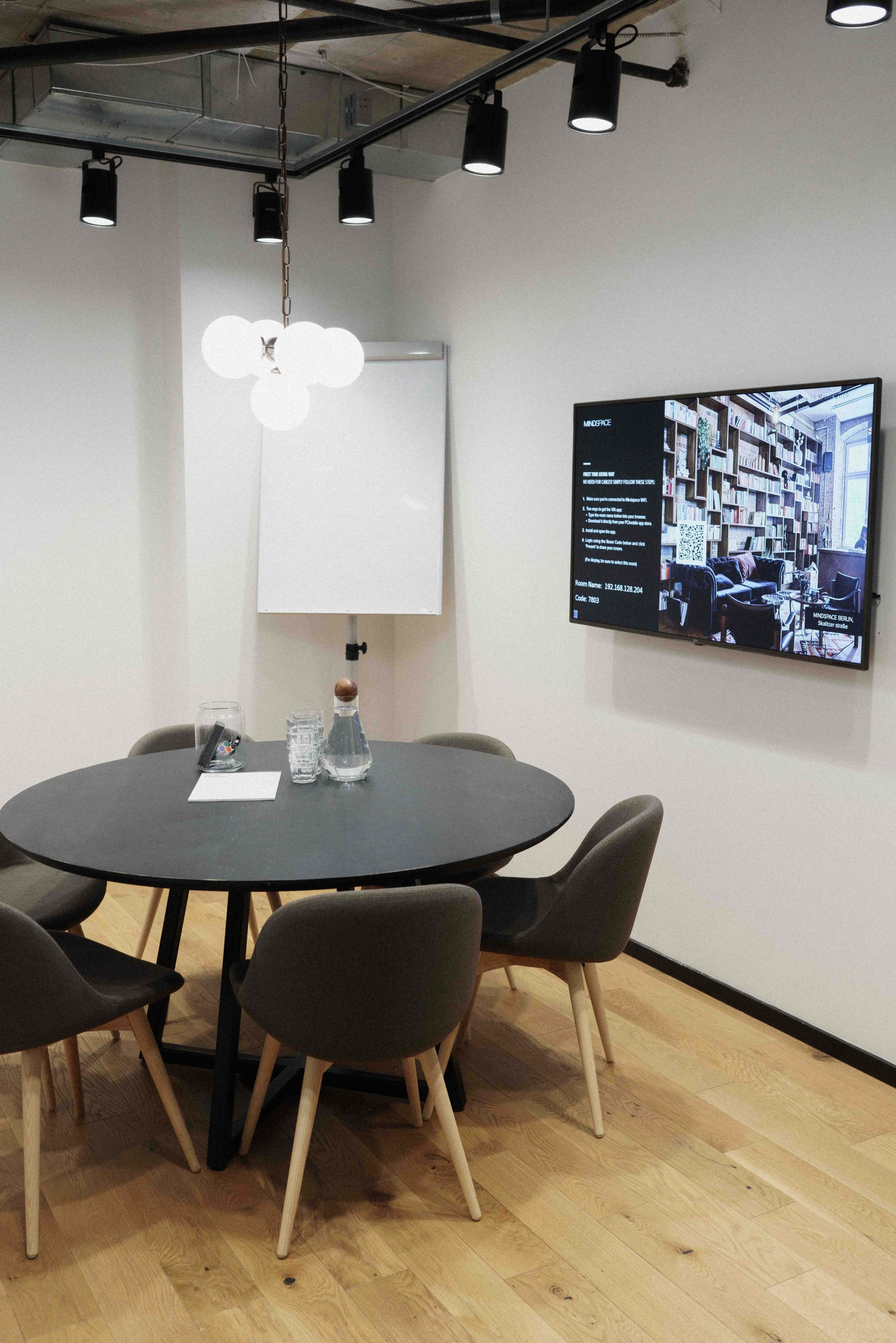 A modern meeting room features a round black table surrounded by four chairs, a flip chart, a water pitcher, and a wall-mounted screen displaying information.