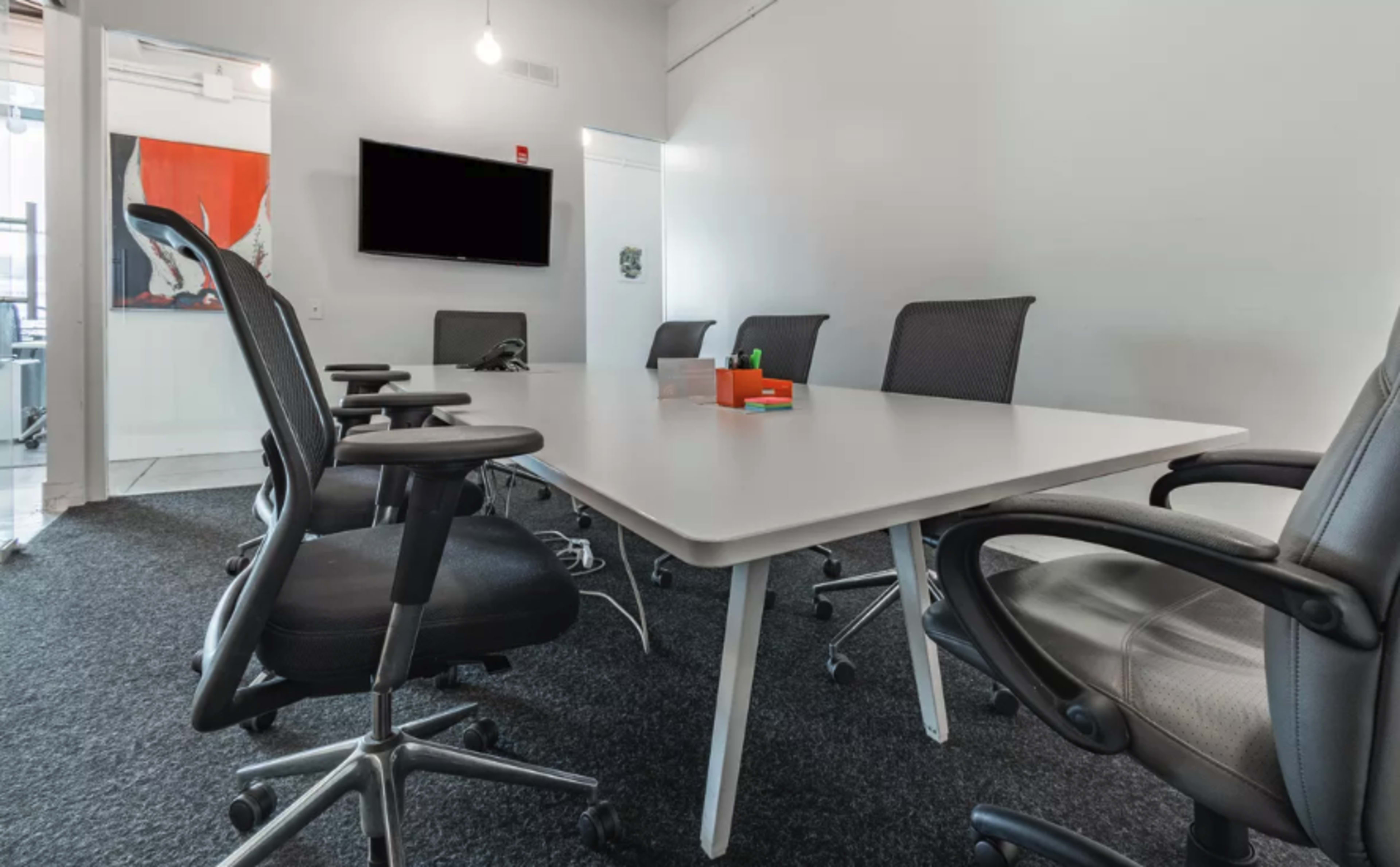 A conference room features a large, rectangular table surrounded by ergonomic office chairs and a wall-mounted screen.