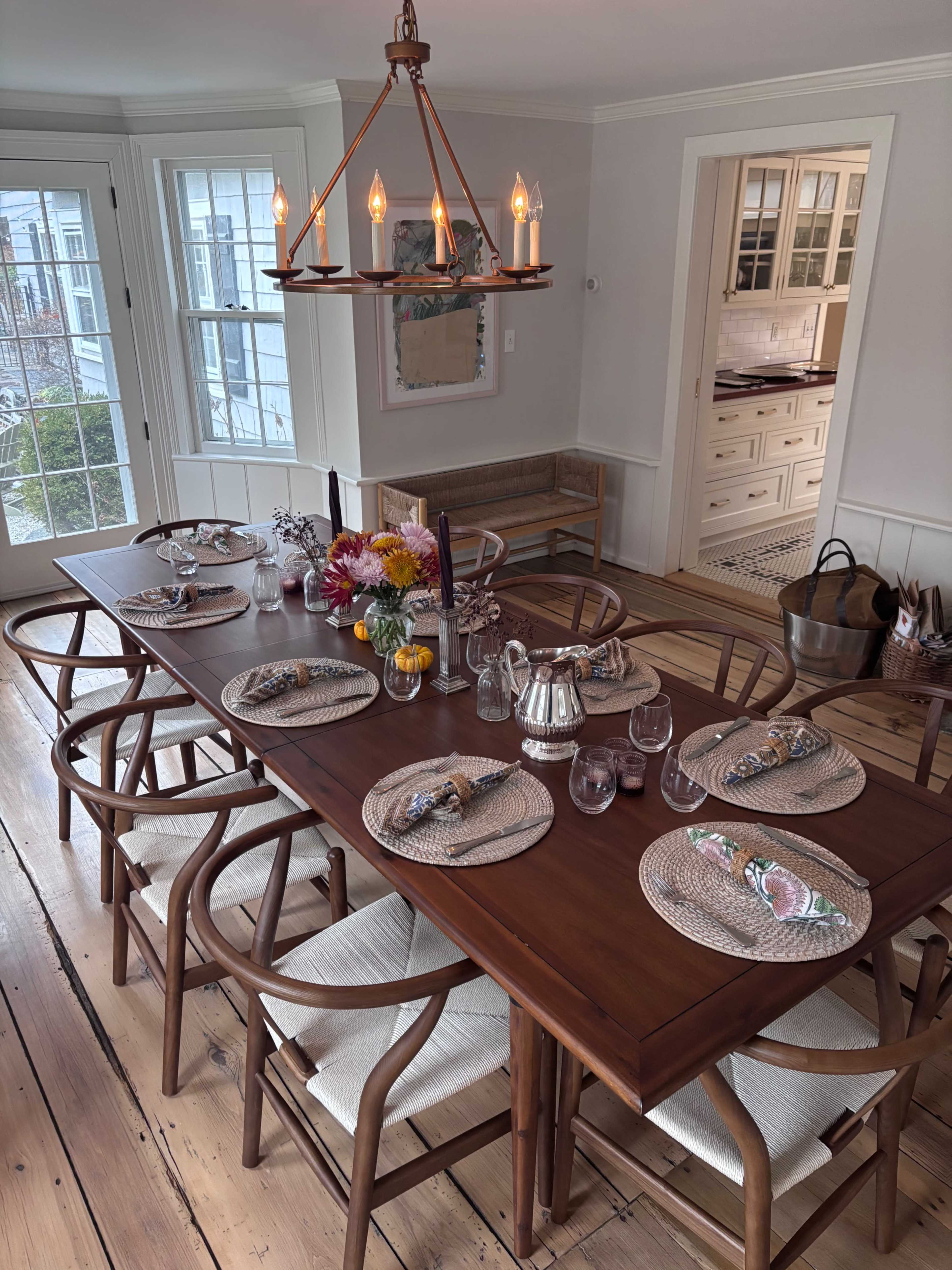 A wooden dining table is set for a meal, featuring plates, glassware, and napkins, with a light fixture above and a view of a kitchen in the background.