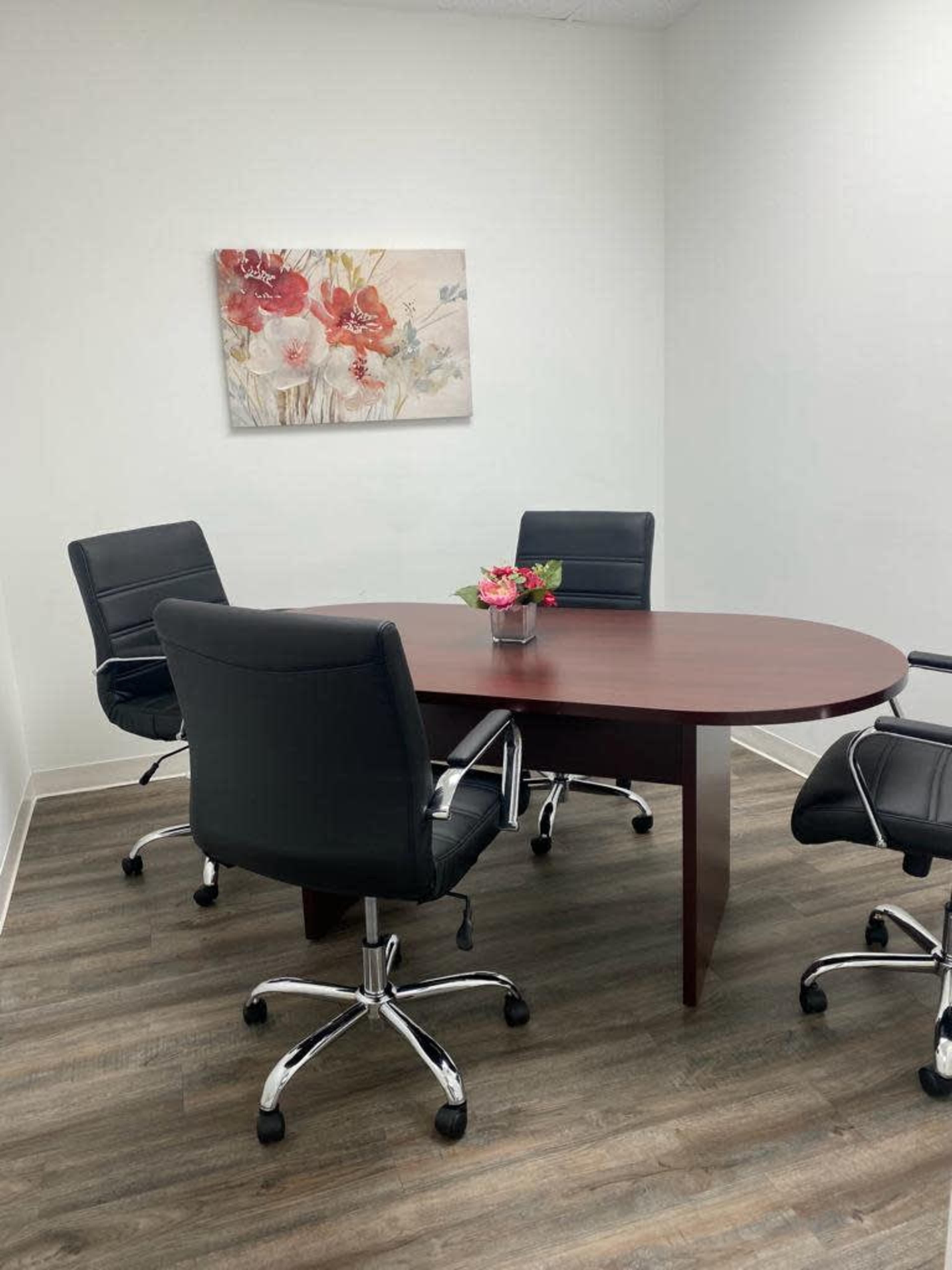A small meeting room features a round wooden table surrounded by four black swivel chairs, with a flower arrangement in the center and a floral painting on the wall.