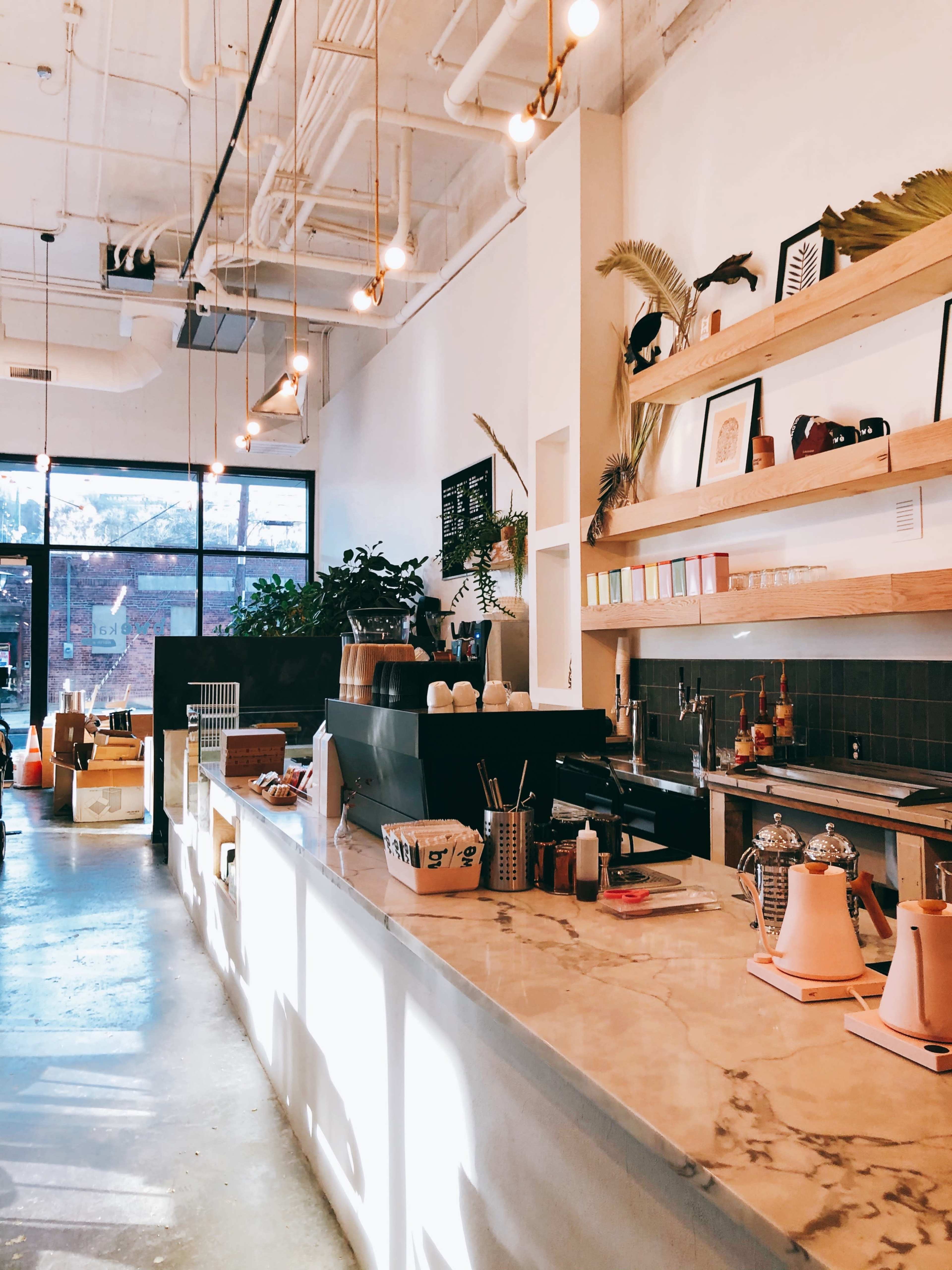 The image shows a bright, modern coffee shop interior with a marble counter, wooden shelves displaying decorative items, and plants in the background.