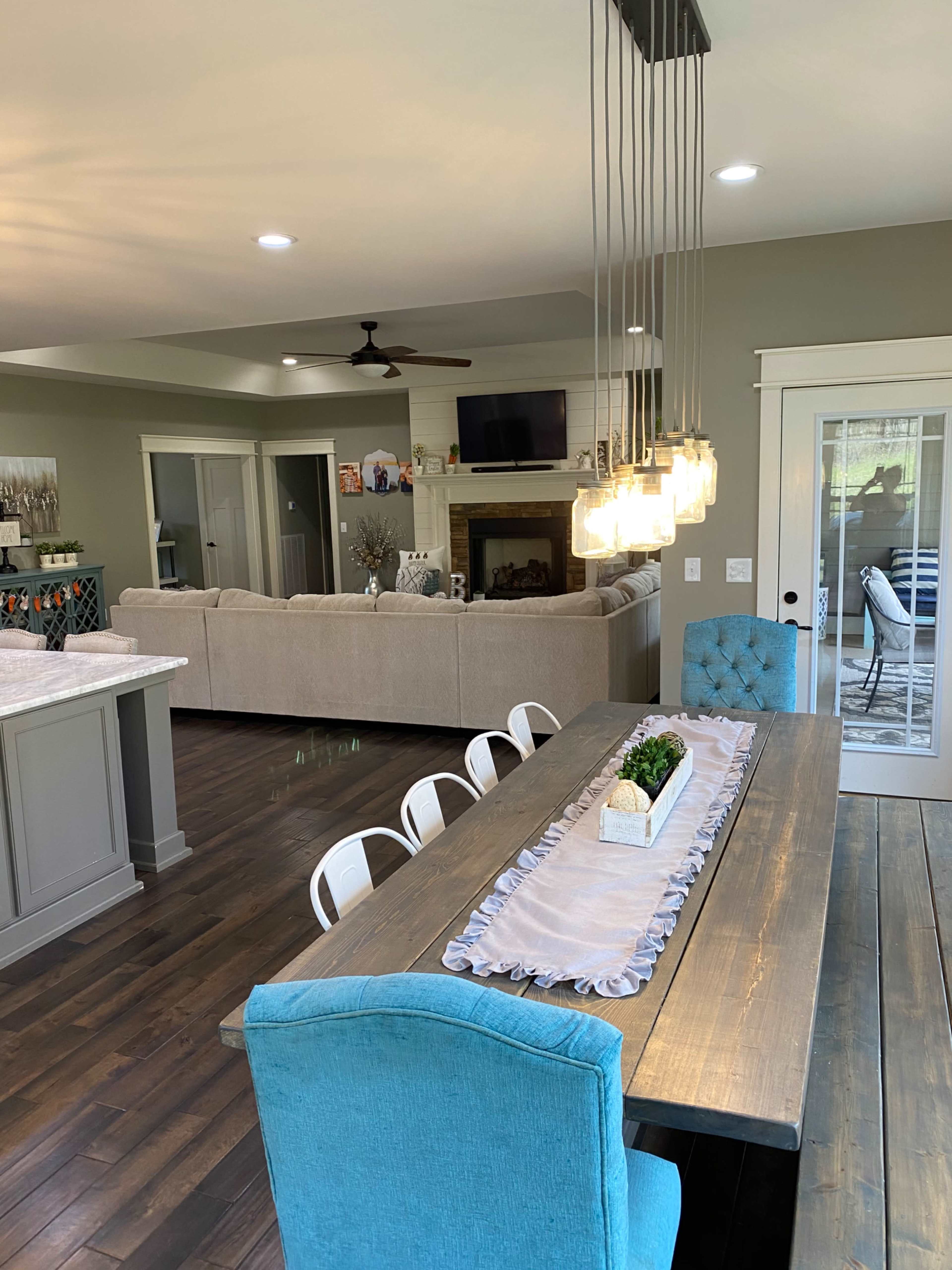 A modern dining area with a long wooden table, blue upholstered chairs, and pendant lighting, overlooking a living room with a fireplace.