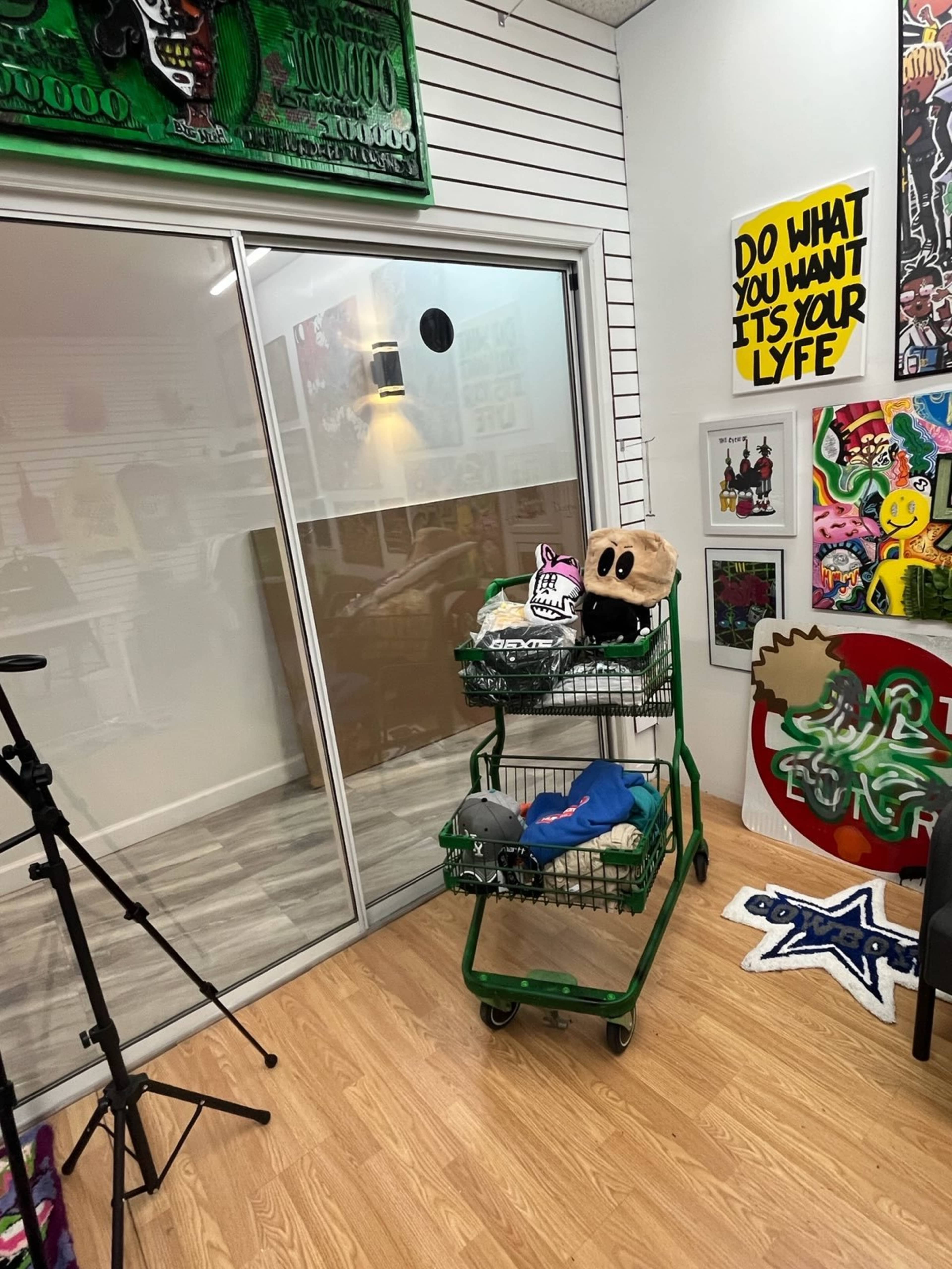 A shopping cart filled with clothing and a plush character sits in a room decorated with colorful artwork and a glass door.