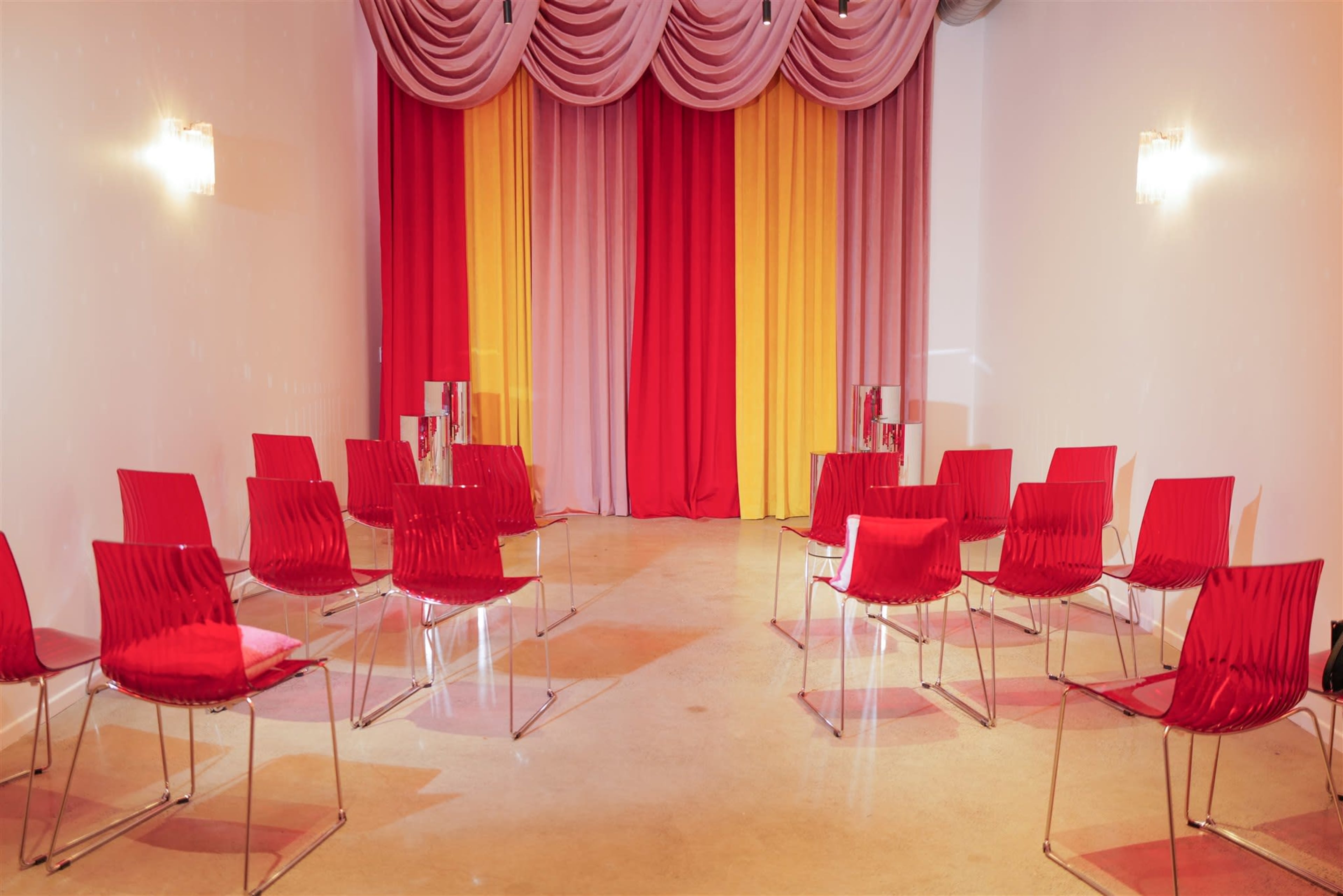 A room set up with several red chairs arranged in a semi-circle facing a backdrop of draped fabric in red, pink, and yellow.