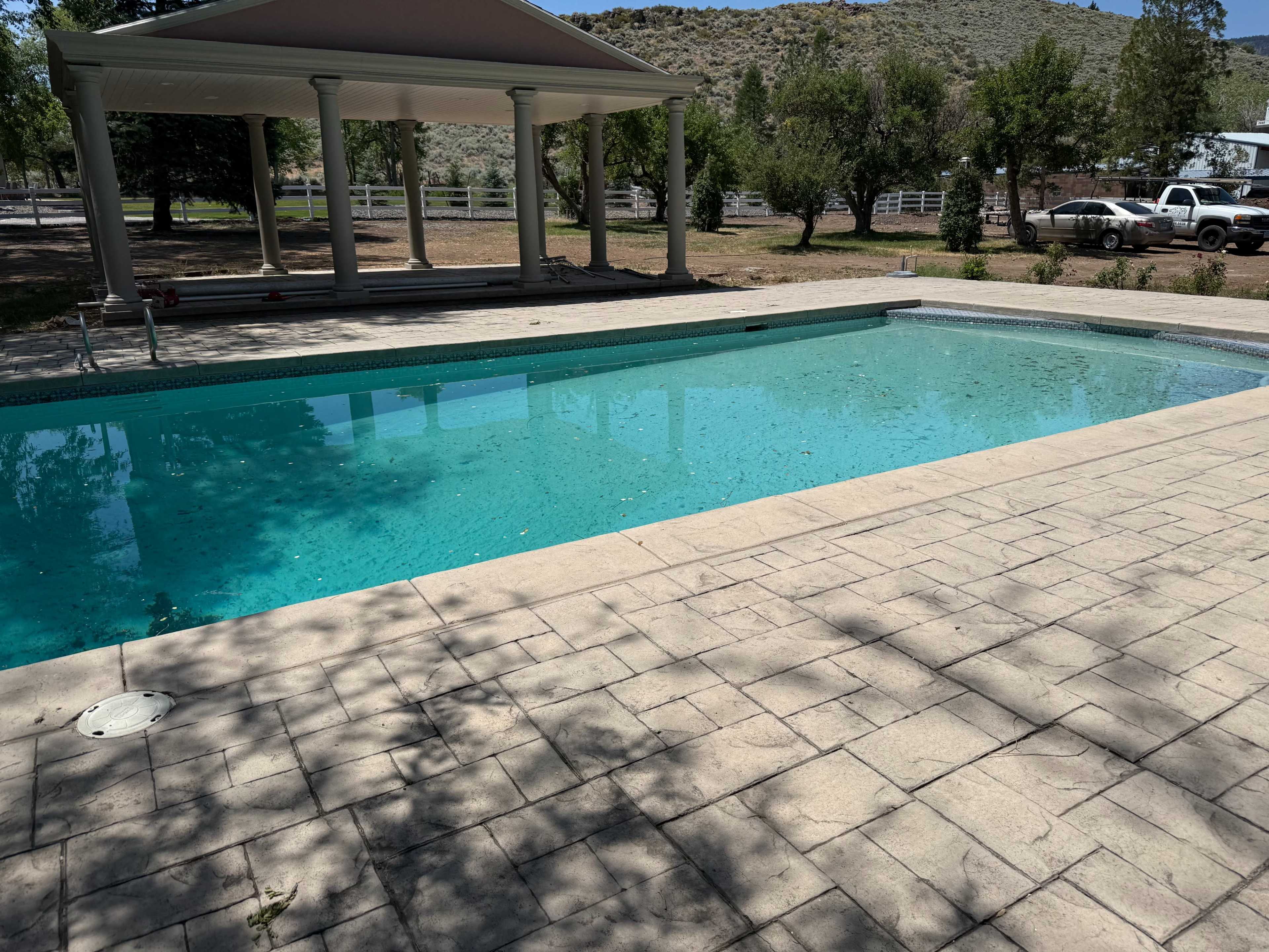 The image shows a rectangular swimming pool surrounded by a stone patio and shaded by adjacent trees.