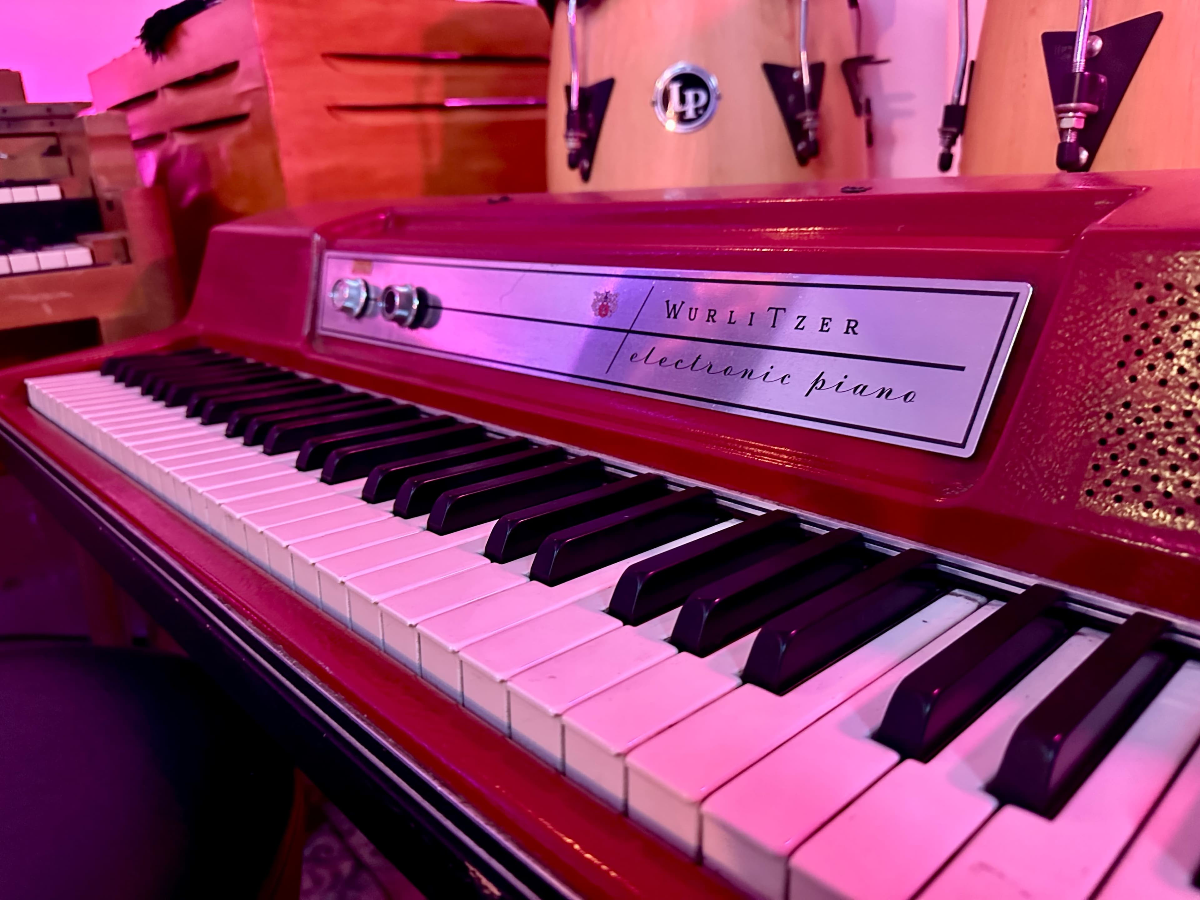 The image shows a close-up of a Wurlitzer electronic piano with a black and white keyboard, set against a backdrop featuring percussion instruments.