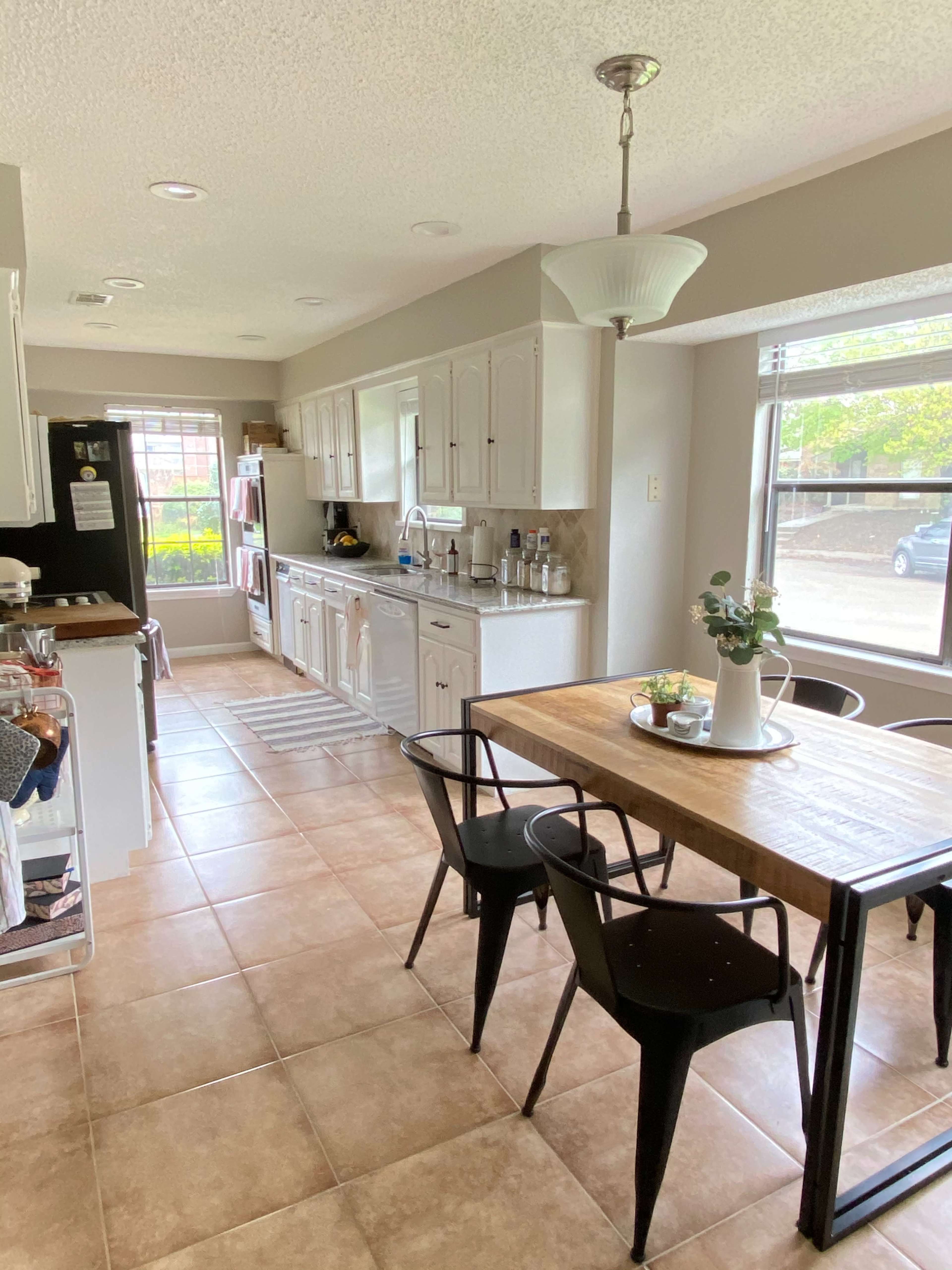 The image shows a modern kitchen with white cabinets, a wooden dining table, and a view of a patio through large windows.
