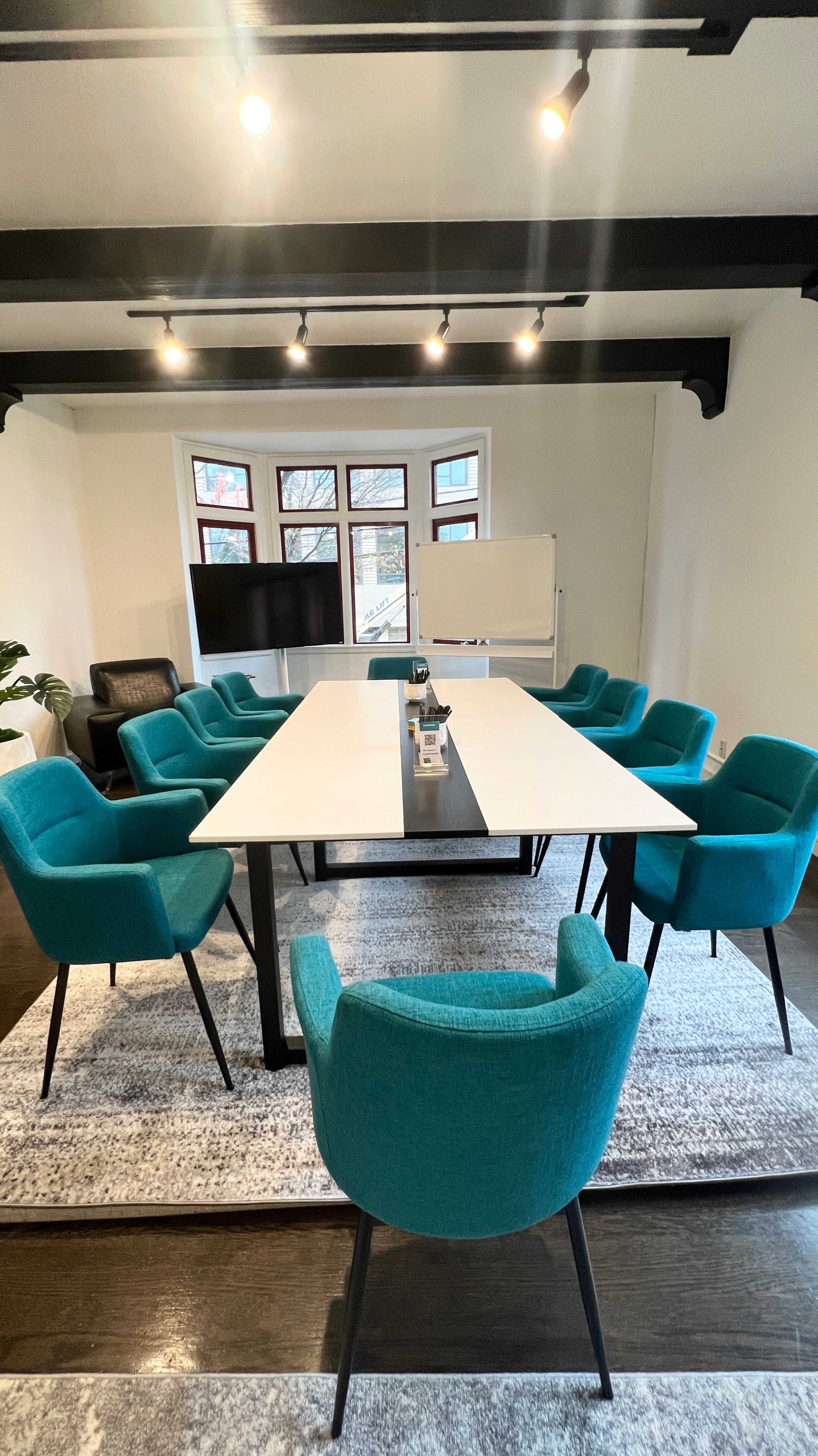 A conference room with a long white table surrounded by ten teal chairs and a large screen at one end.