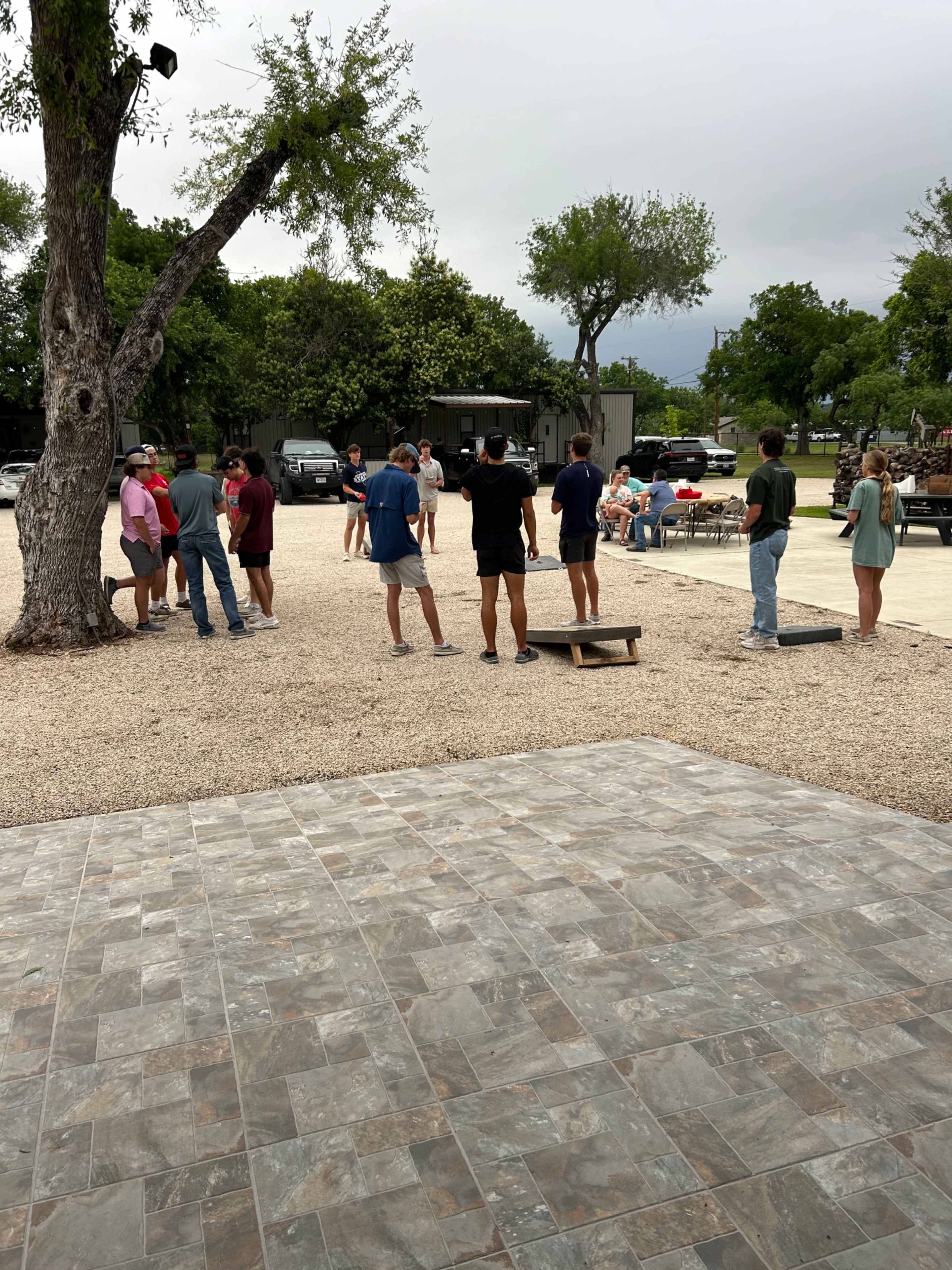 A group of people stands in a gravel area with a tree, facing each other while others are seated nearby.