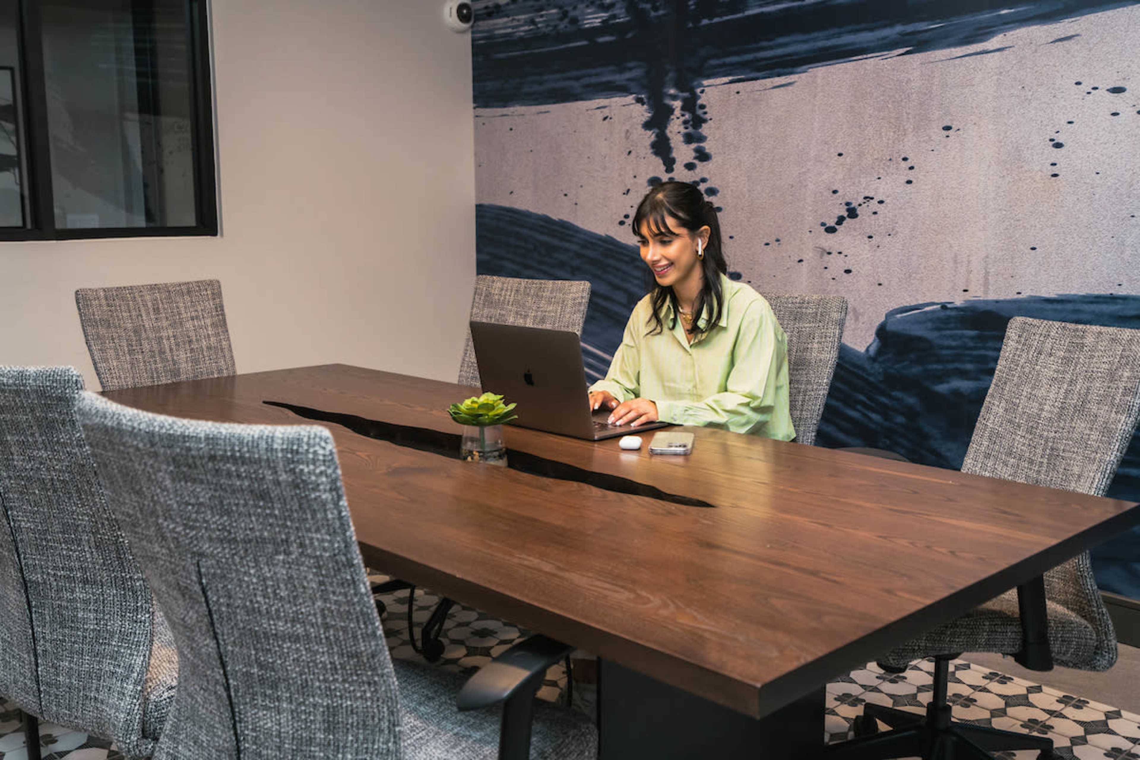 A woman sits at a conference table using a laptop in a modern meeting room with a textured wall mural.