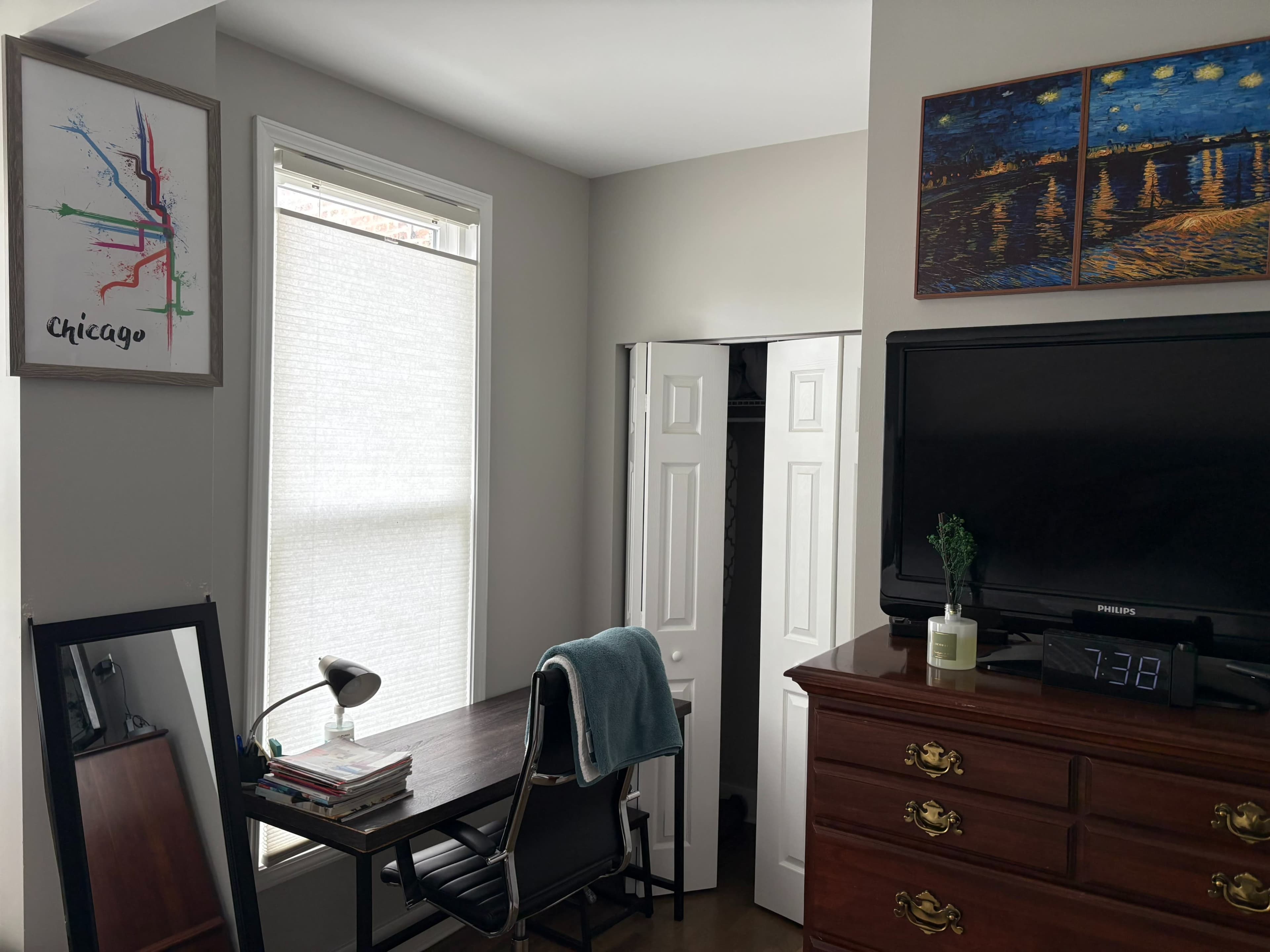The image shows a small room with a desk, chair, lamp, and a television, along with a closed closet and a window covered by a light shade.