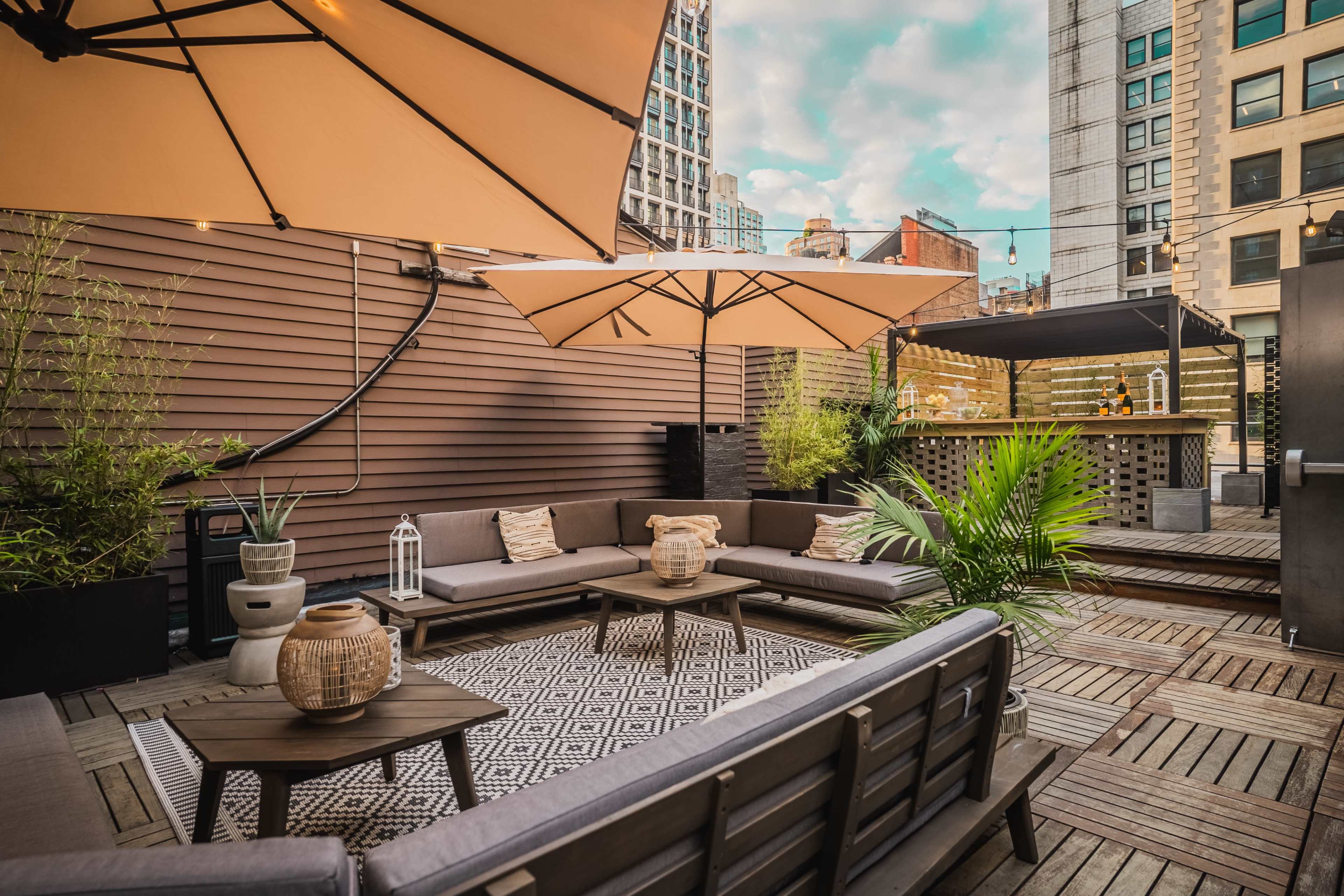A rooftop terrace with lounge seating, large umbrellas, and a wooden bar surrounded by greenery and city buildings.
