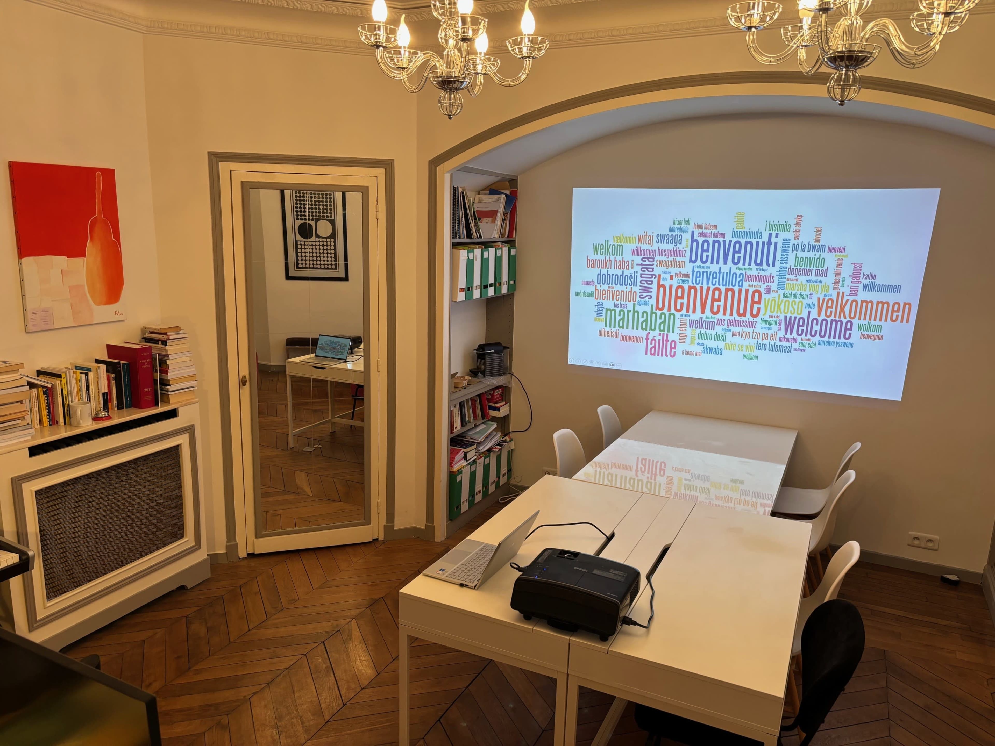 A bright meeting room features a large projection of a multicolored word cloud displaying greetings in various languages on one wall, with a modern white table and chairs in the center.