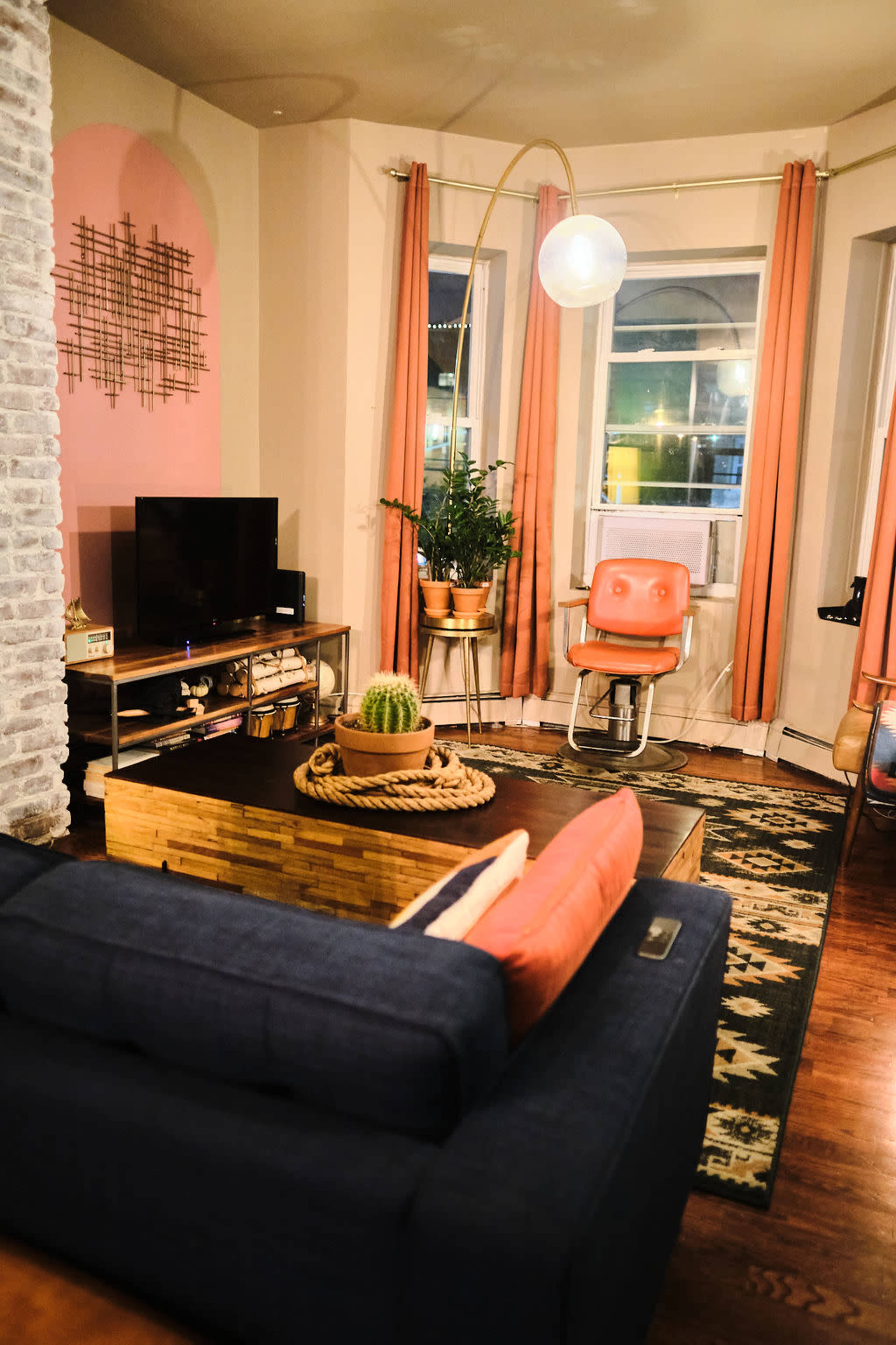A cozy living room with a blue sofa, a wooden coffee table centered with a cactus, a television on a stand, and a tall lamp beside a retro-style chair.