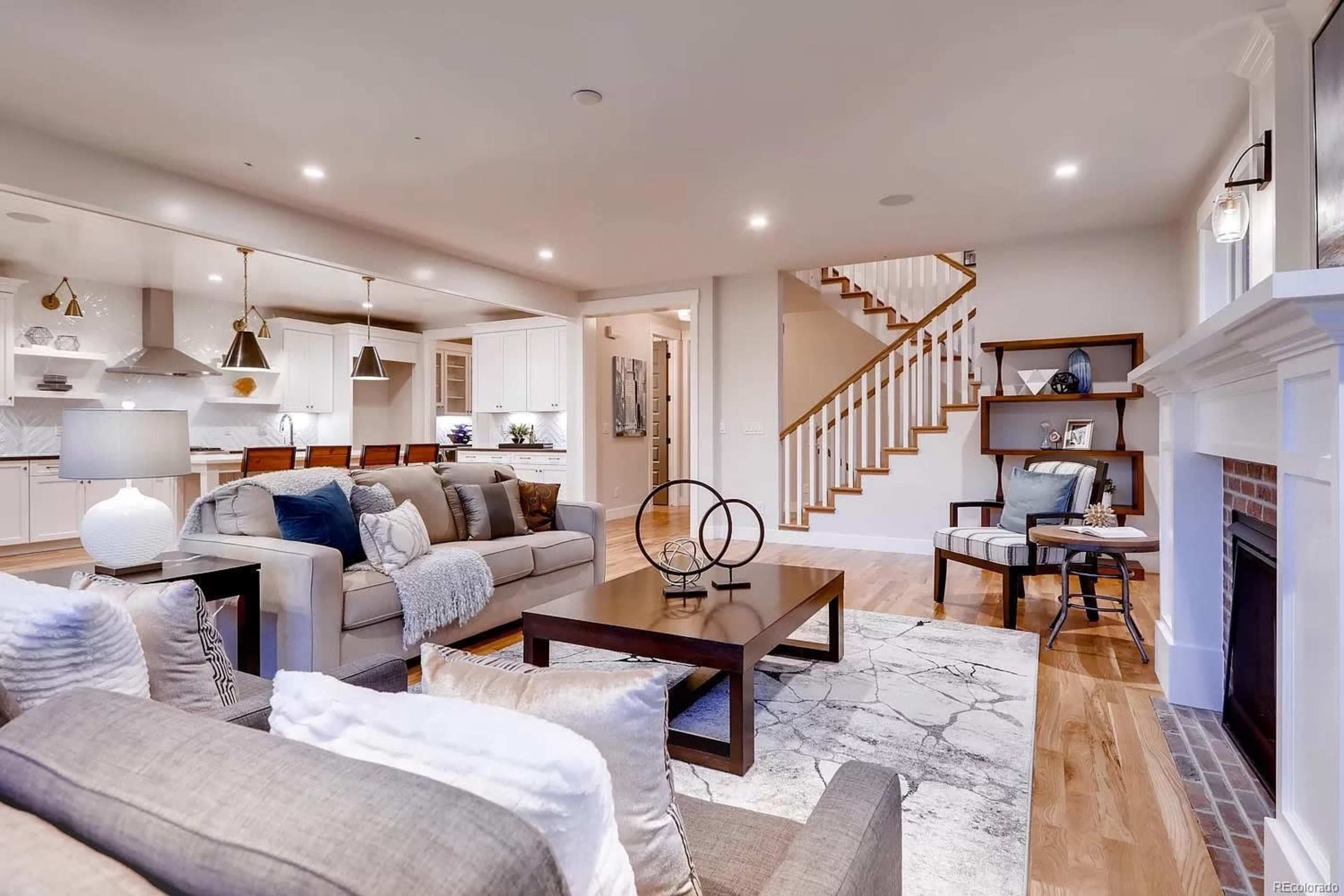 The image shows a bright, modern living room connected to a kitchen, featuring a large coffee table, a sofa with decorative cushions, and a staircase in the background.