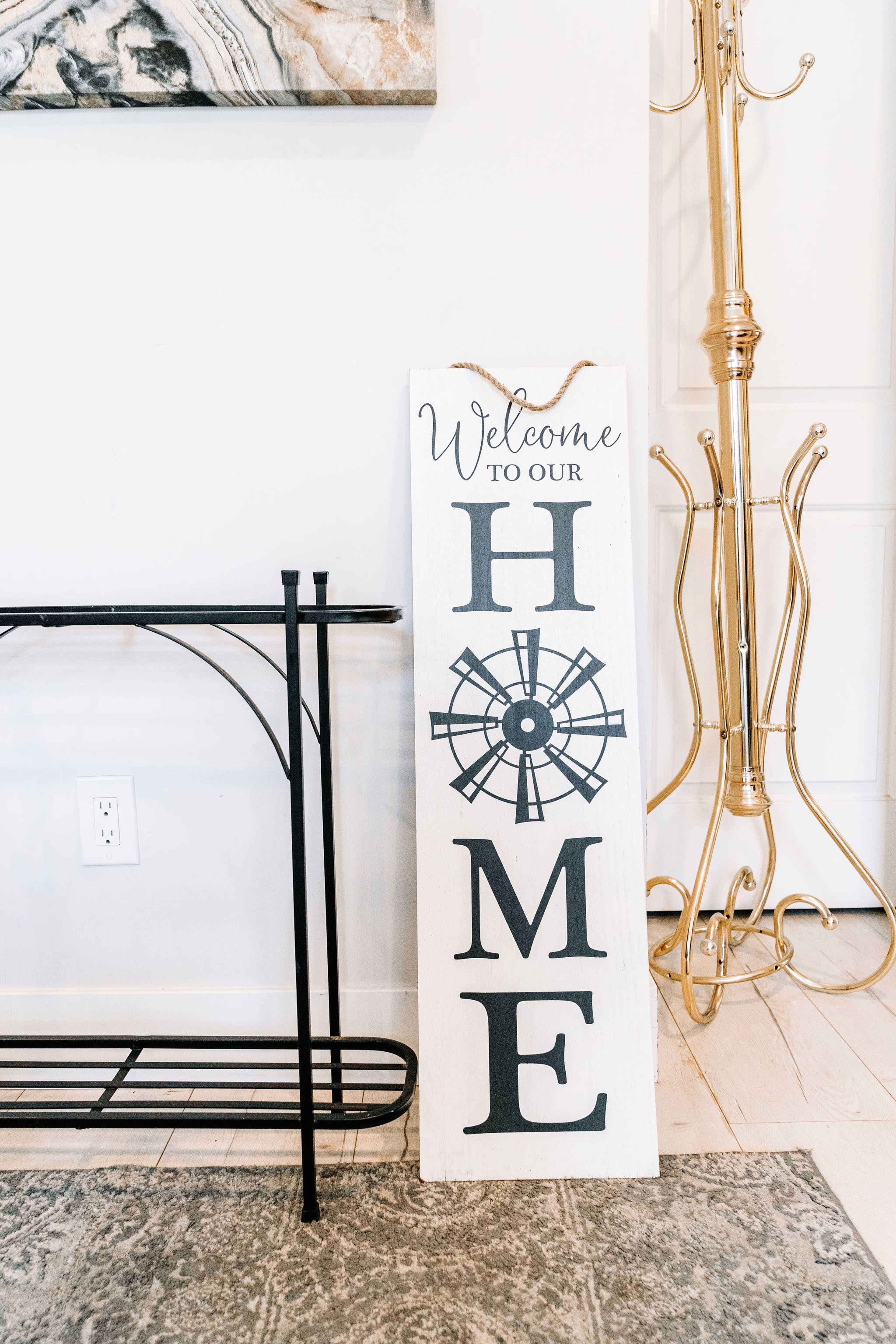 A wooden sign that reads "Welcome to Our HOME" with a decorative design, placed next to a metal coat rack in a well-lit interior space.