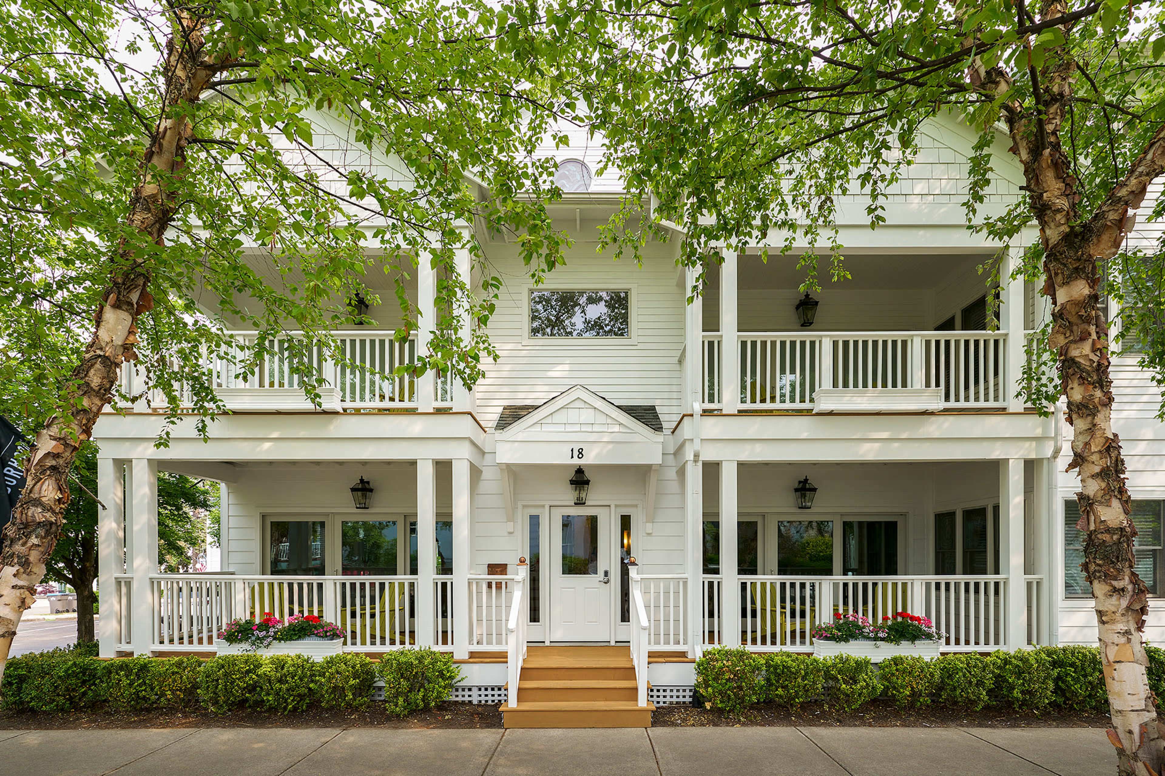A white two-story house with multiple balconies, framed by trees, features a central entrance labeled "18" and flower beds on either side.