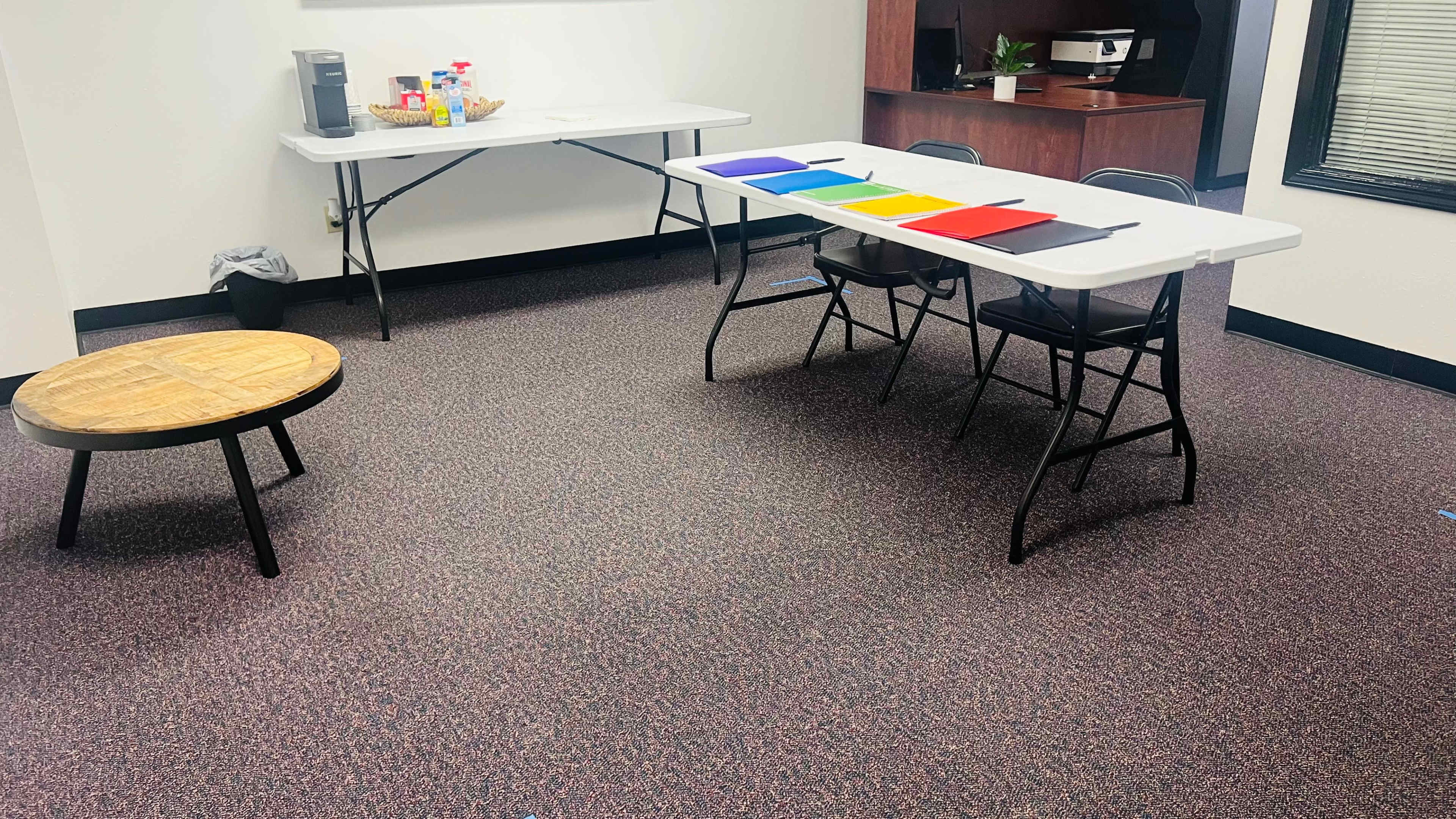 A room with a small round table, two folding tables with colored folders arranged in a row, and a snack station in the background.