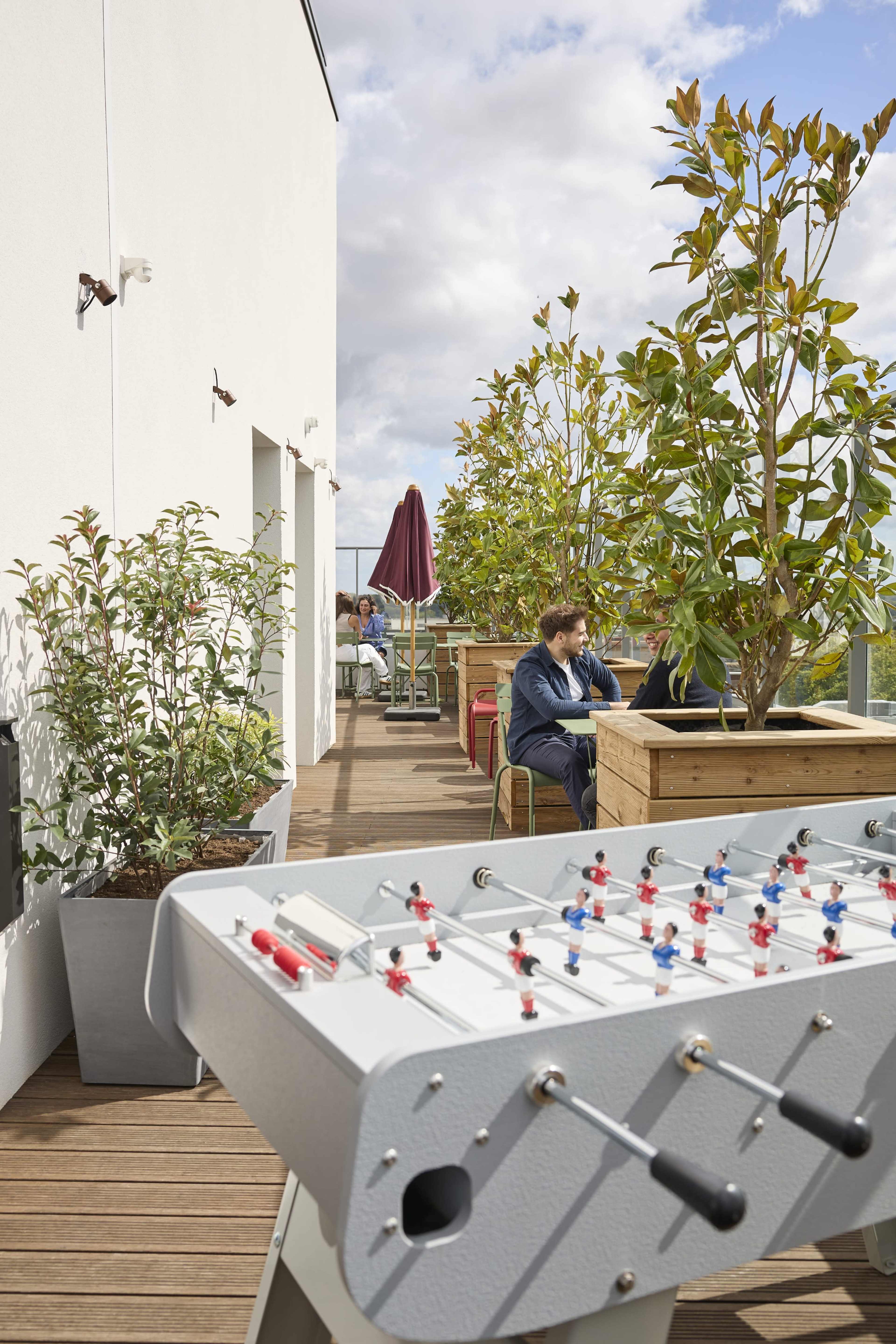 A rooftop terrace features a foosball table, green plants in pots, and a person working on a laptop at a nearby table.