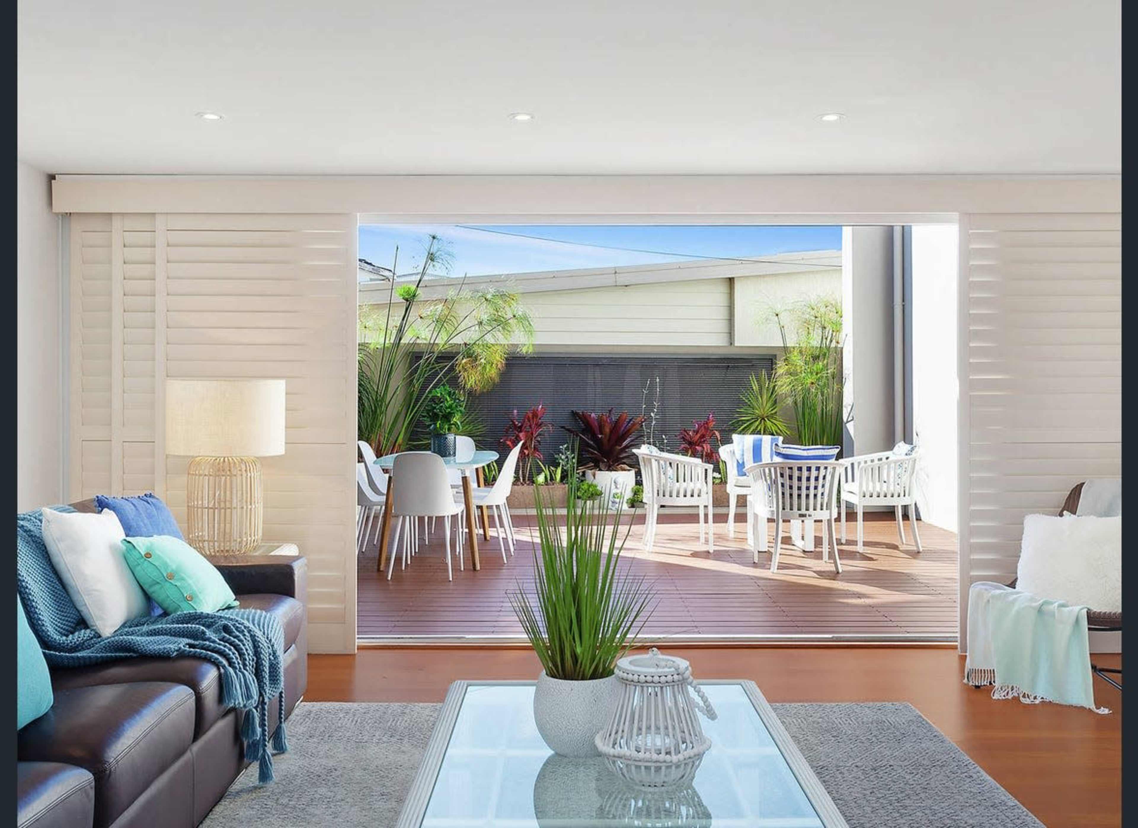 A modern living room features a sliding door that opens to a patio with outdoor seating and lush greenery.