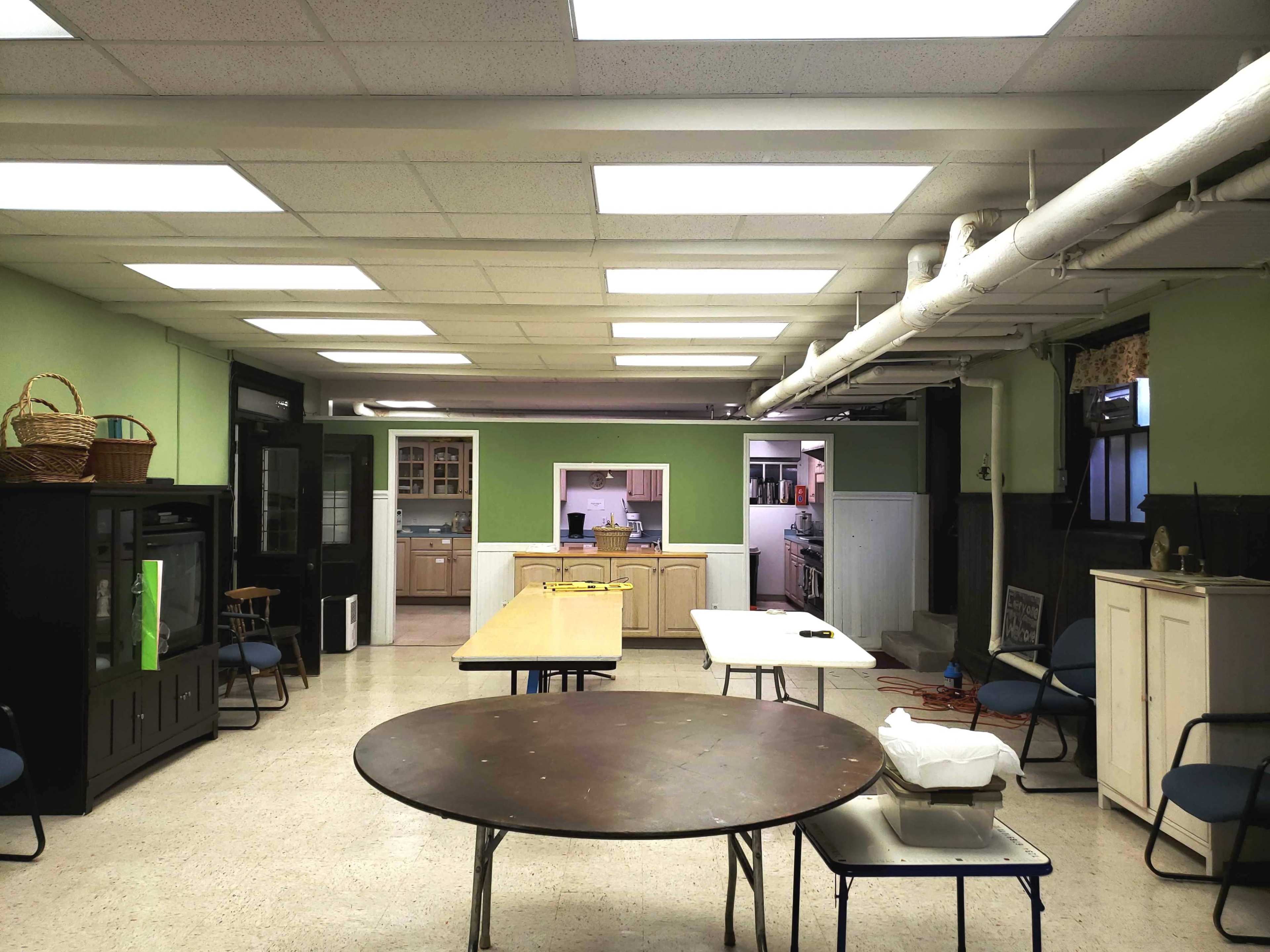 A spacious room with a kitchen area in the background, featuring several tables and chairs arranged throughout.