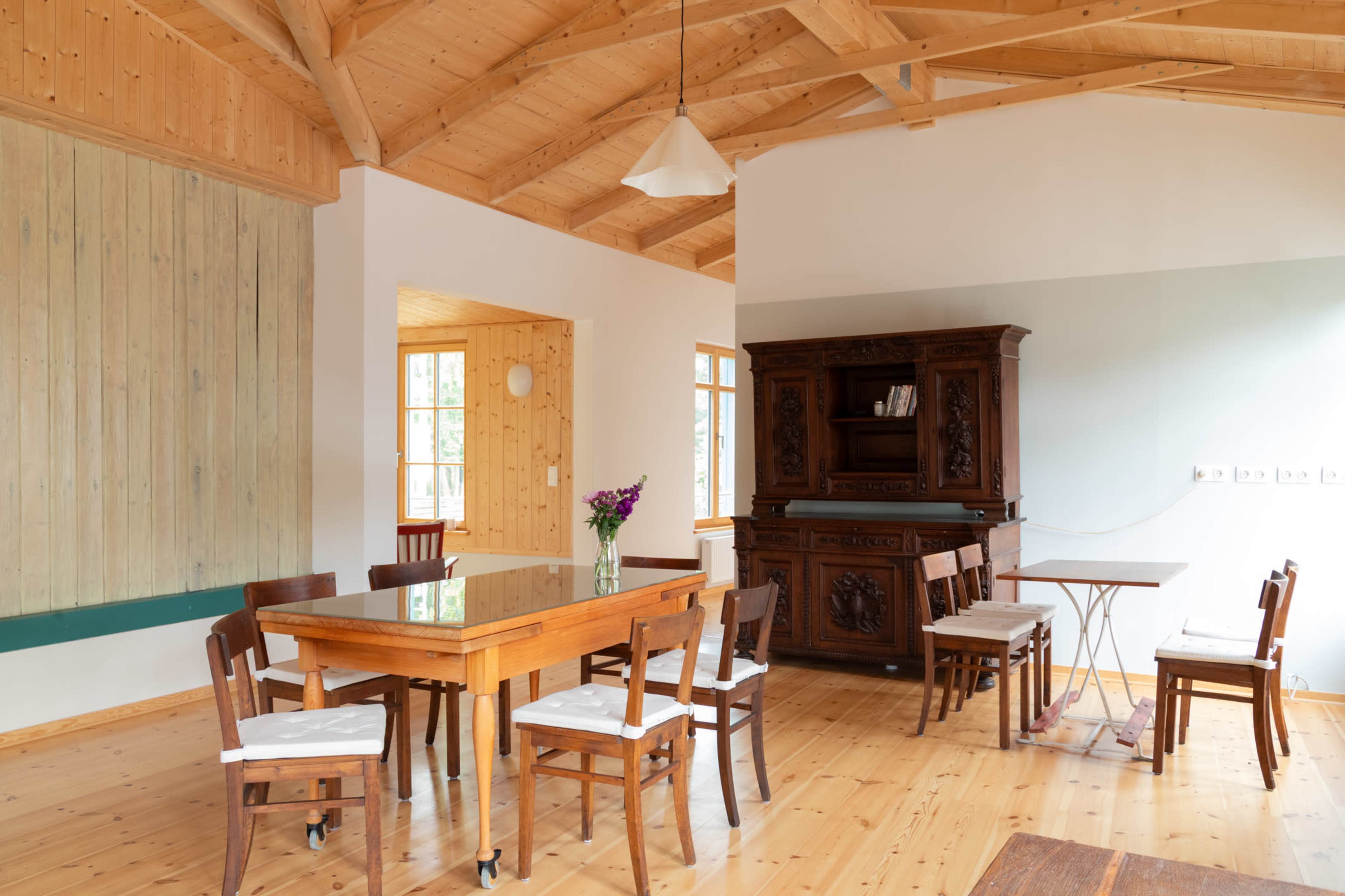 The image shows a spacious room with wooden walls and ceiling, featuring a large table in the center, surrounded by chairs, and a dark wooden cabinet against the wall.