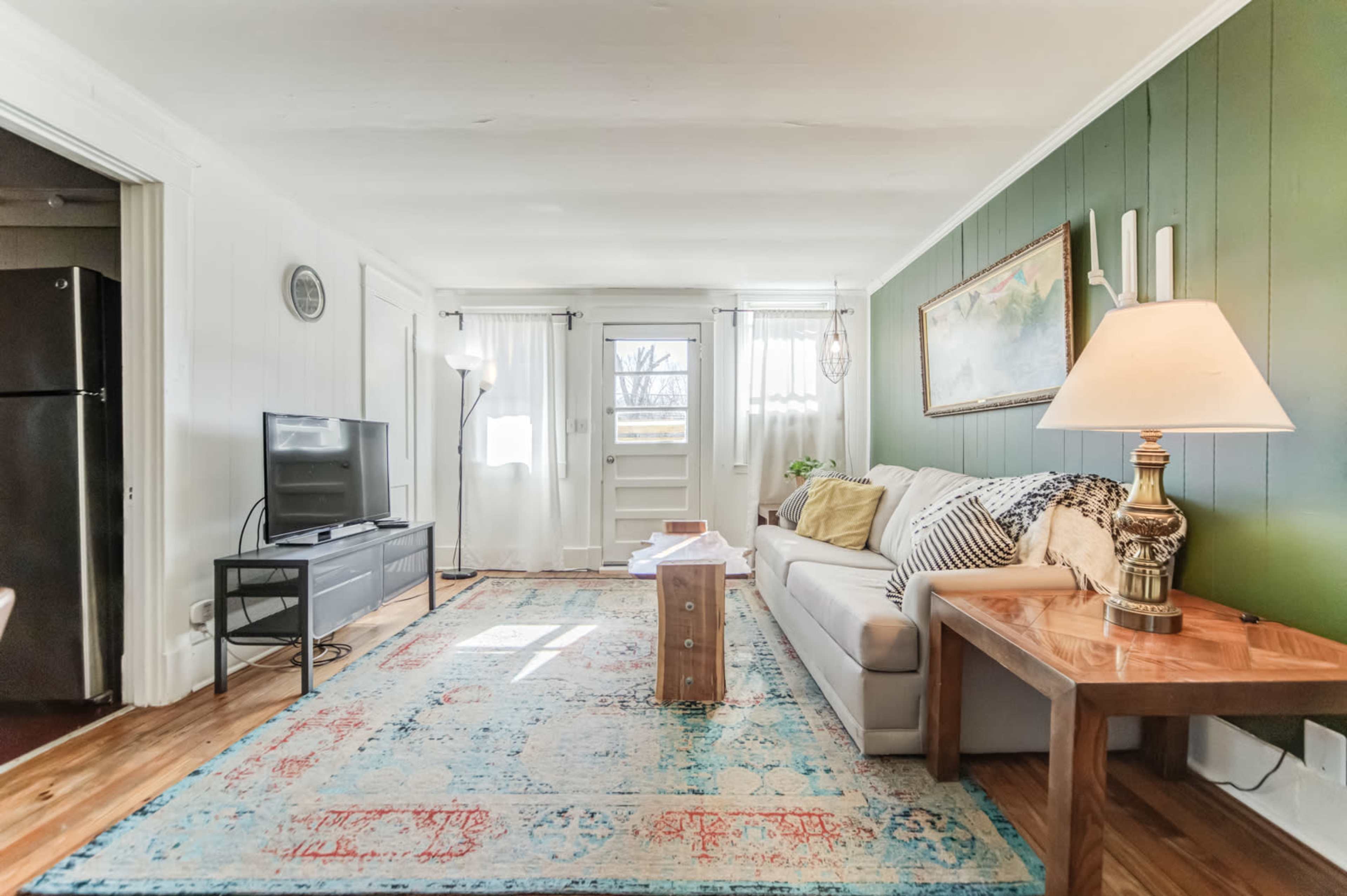 The living room features a sofa, a coffee table, a television, and large windows, with green accent walls and a decorative rug.