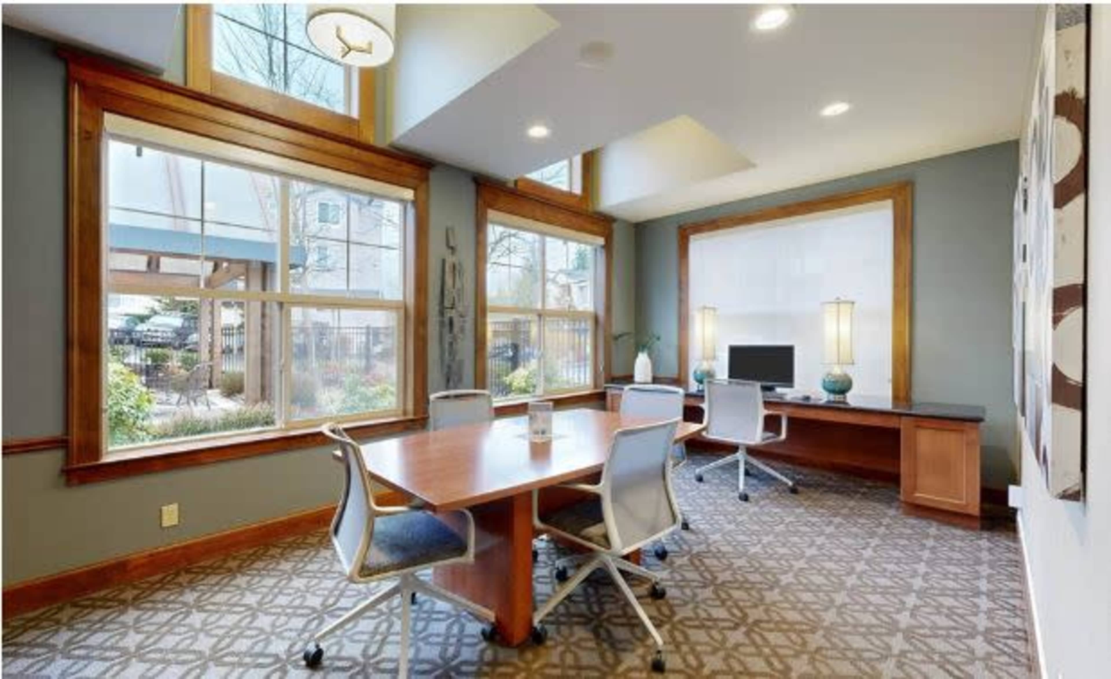 A brightly lit conference room with a wooden table and four chairs, large windows overlooking a garden, and a computer setup on a side desk.