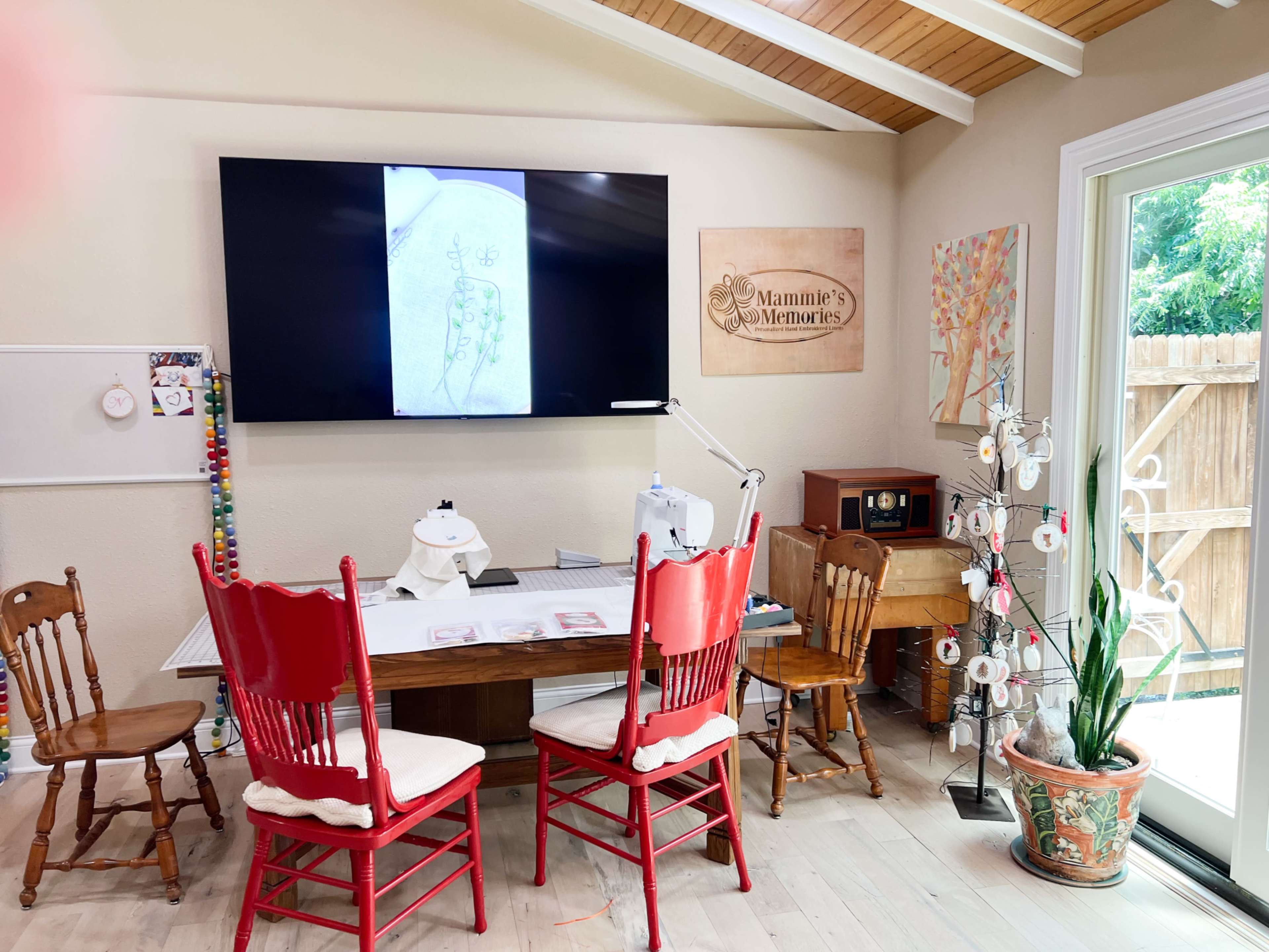A craft room with a large television displaying an illustration, a central table surrounded by red chairs, and a decorative plant in a pot.