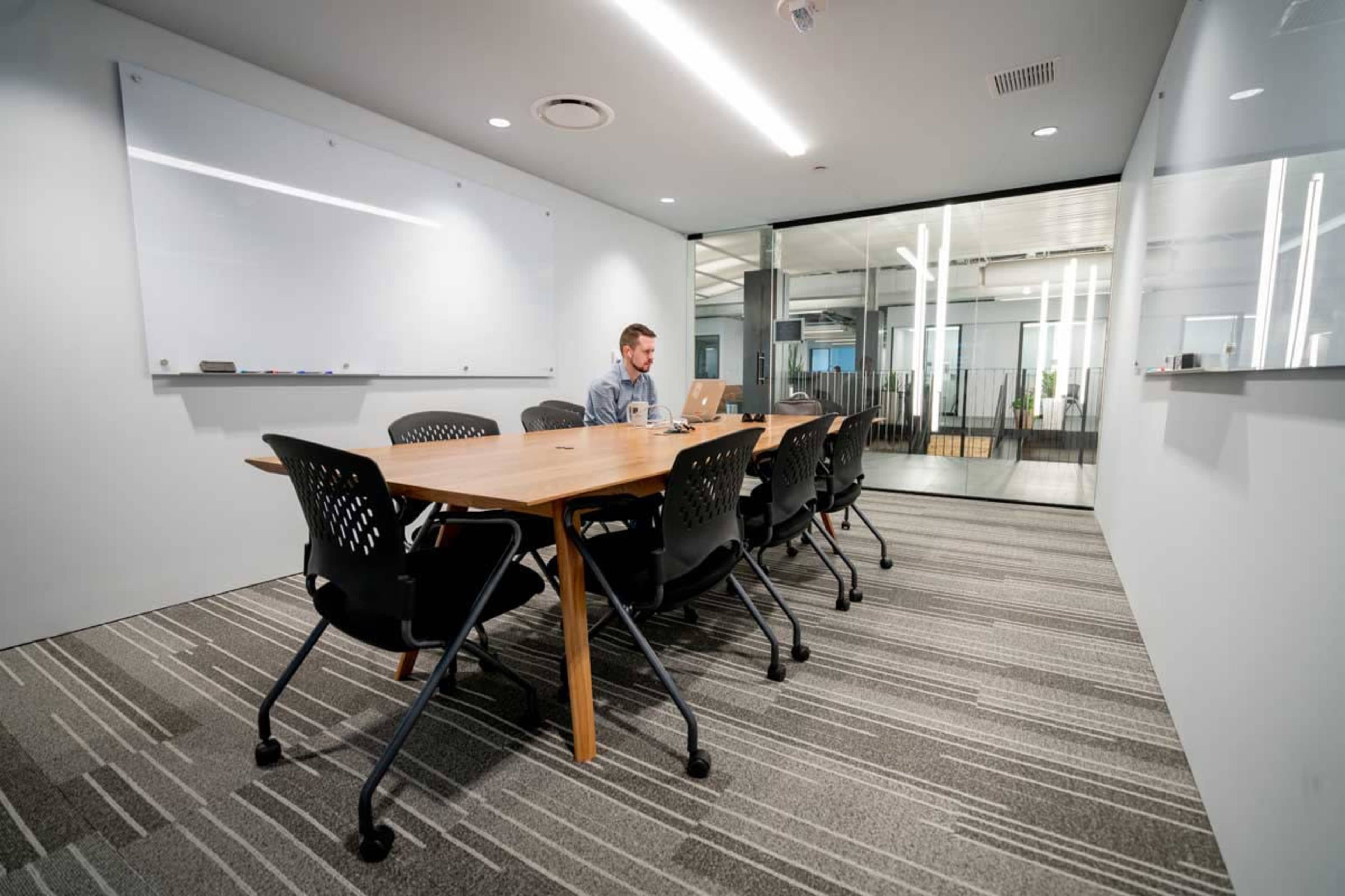 A man sits at a long wooden table in a modern conference room, working on a laptop while surrounded by empty chairs.