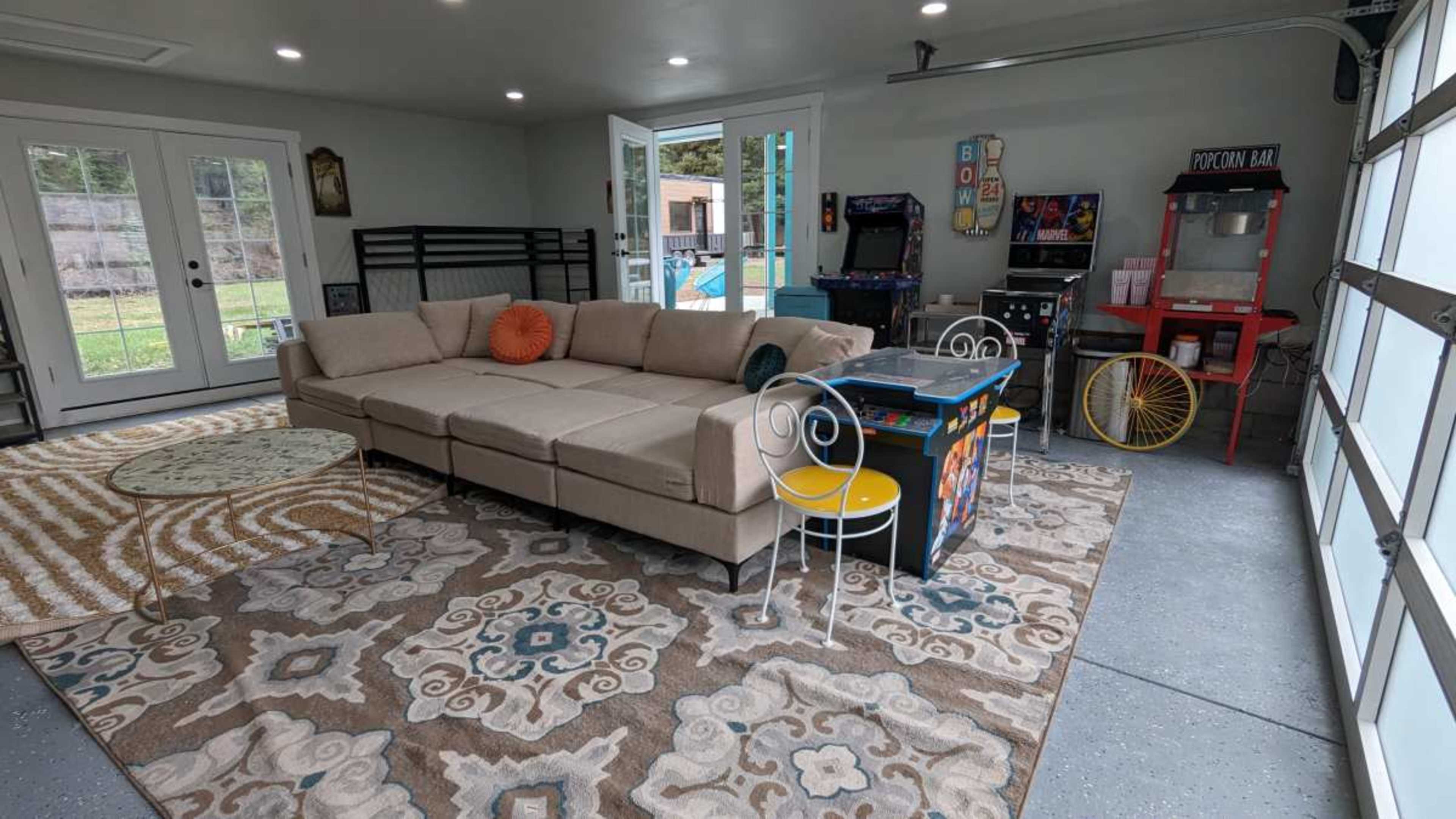 The image shows a spacious game room with a sectional couch, a rug, arcade machines, a popcorn machine, and large windows overlooking a pool area.