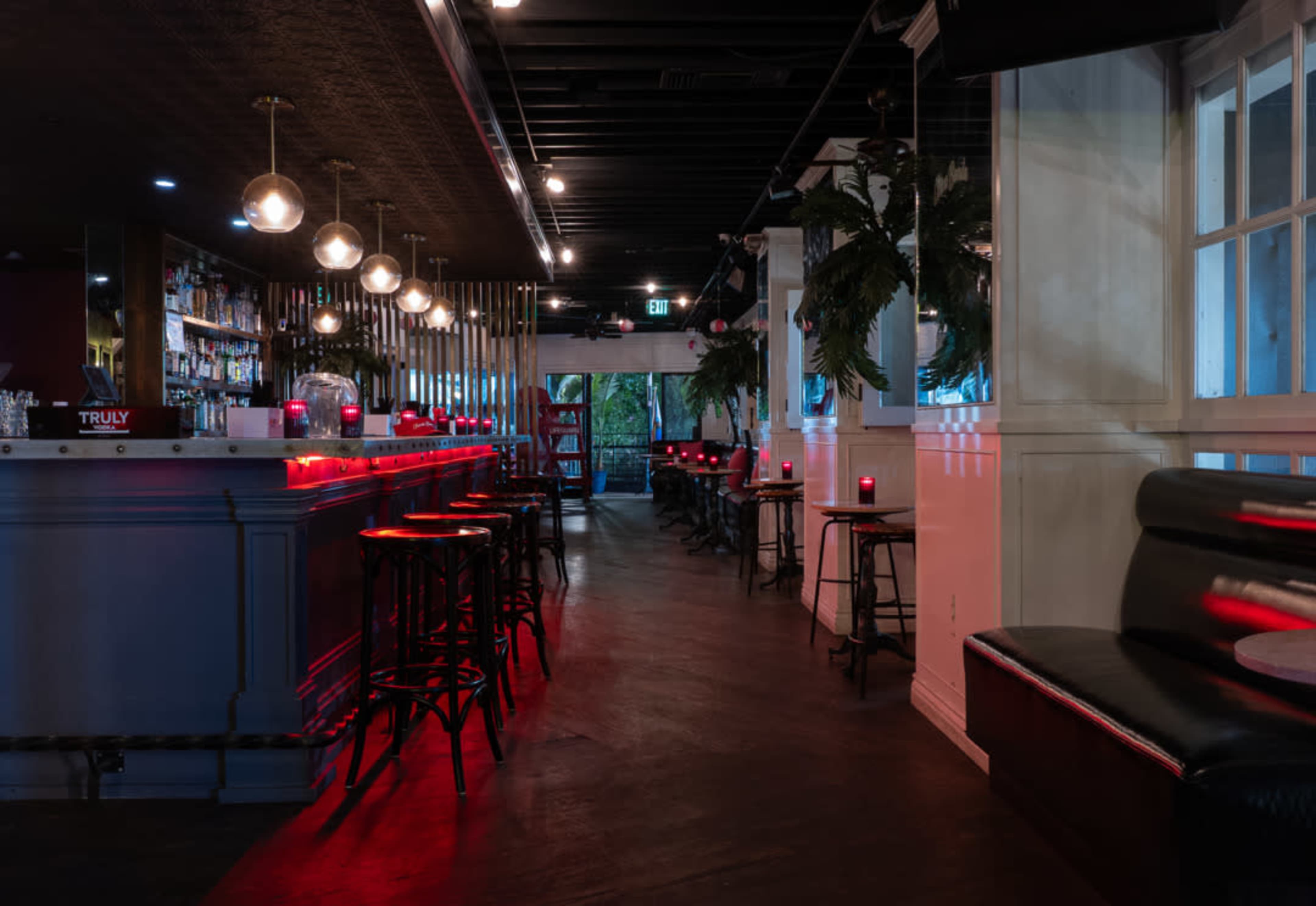 The image shows a dimly lit bar with a long countertop and several high stools, accented by red lighting and decorative plants.
