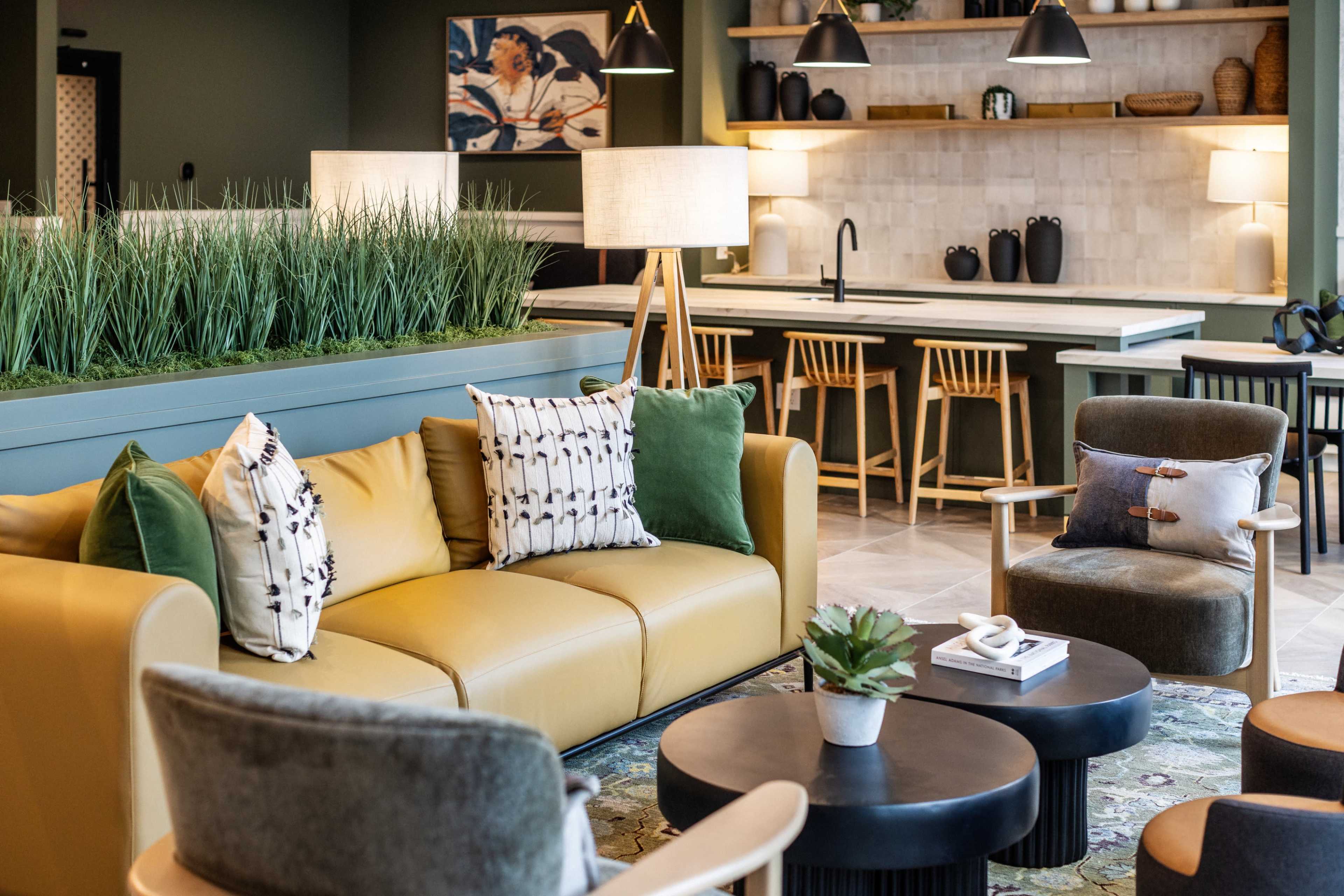 The image shows a modern lounge area with a yellow couch, patterned pillows, circular coffee tables, and a kitchen space with bar stools in the background.