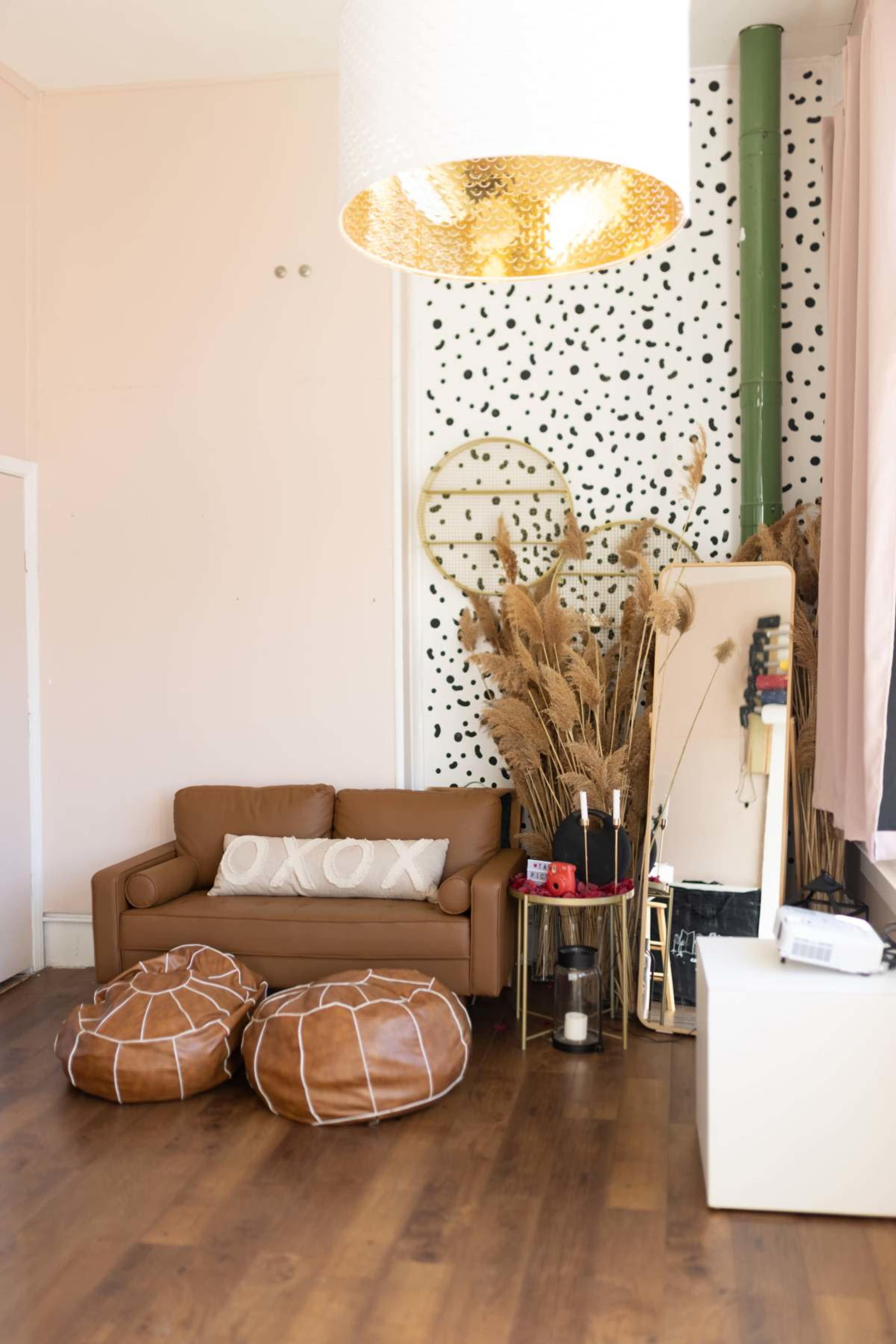 The image shows a modern living room featuring a brown leather sofa, two round brown bean bags, a decorative mirror, and dried plants against a polka-dotted wall.