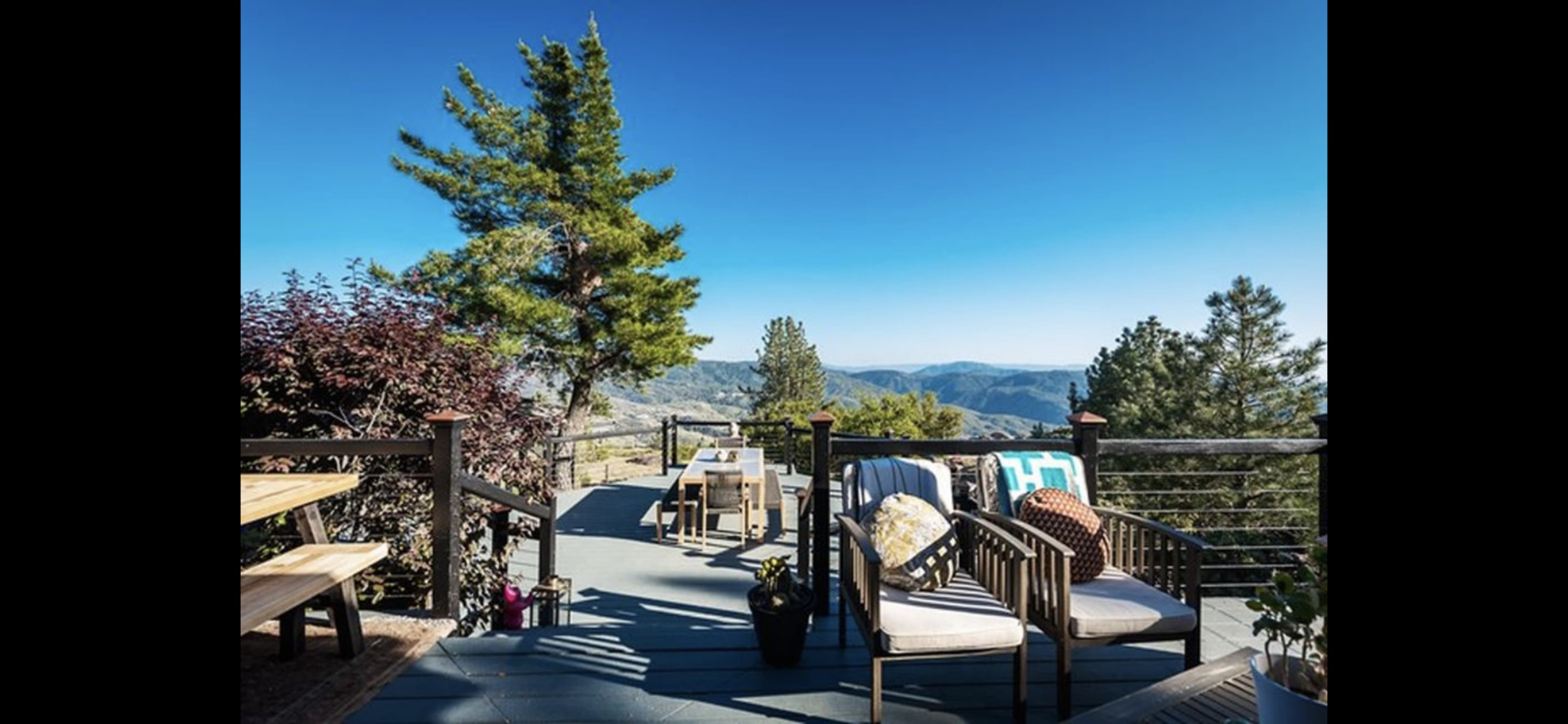 A terrace overlooks a mountainous landscape, featuring wooden seating and potted plants under clear blue skies.