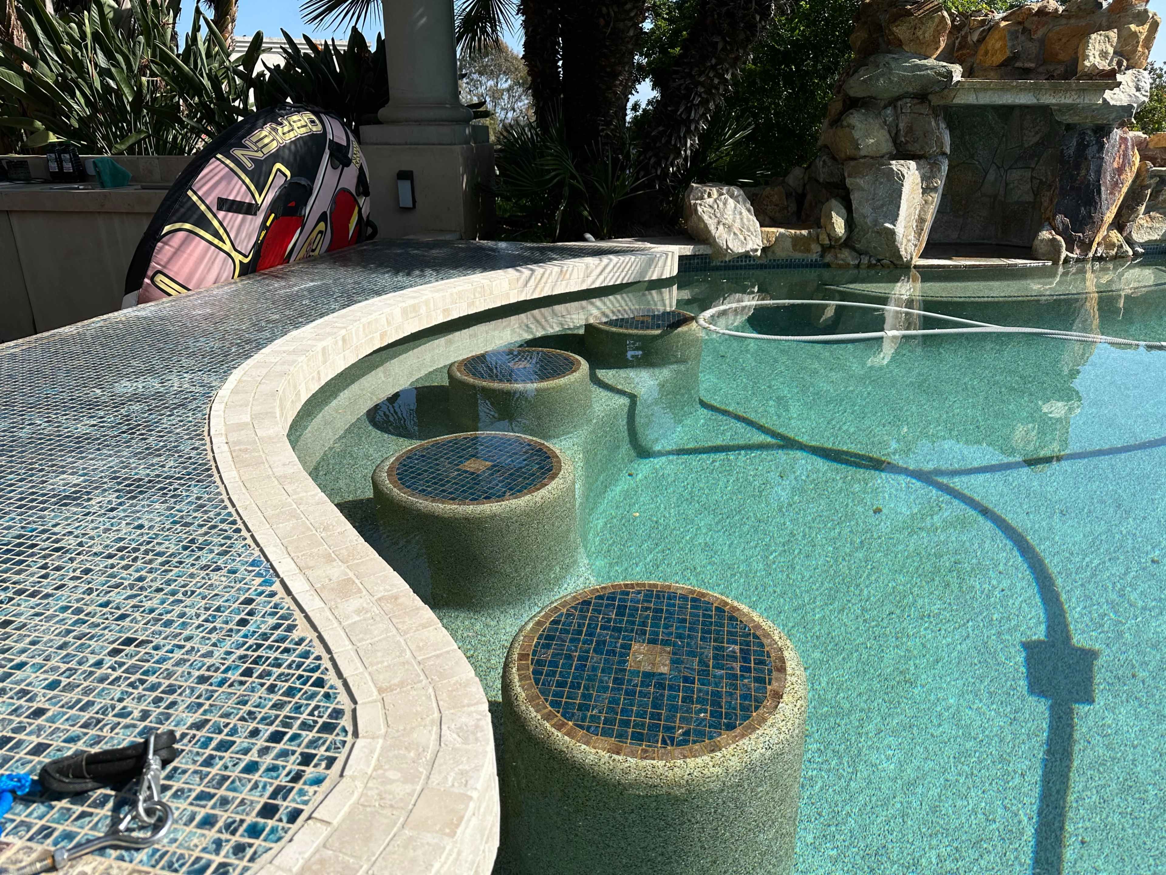 The image shows a swimming pool with circular stone steps partially submerged in clear blue water, surrounded by palm trees and a rock formation.
