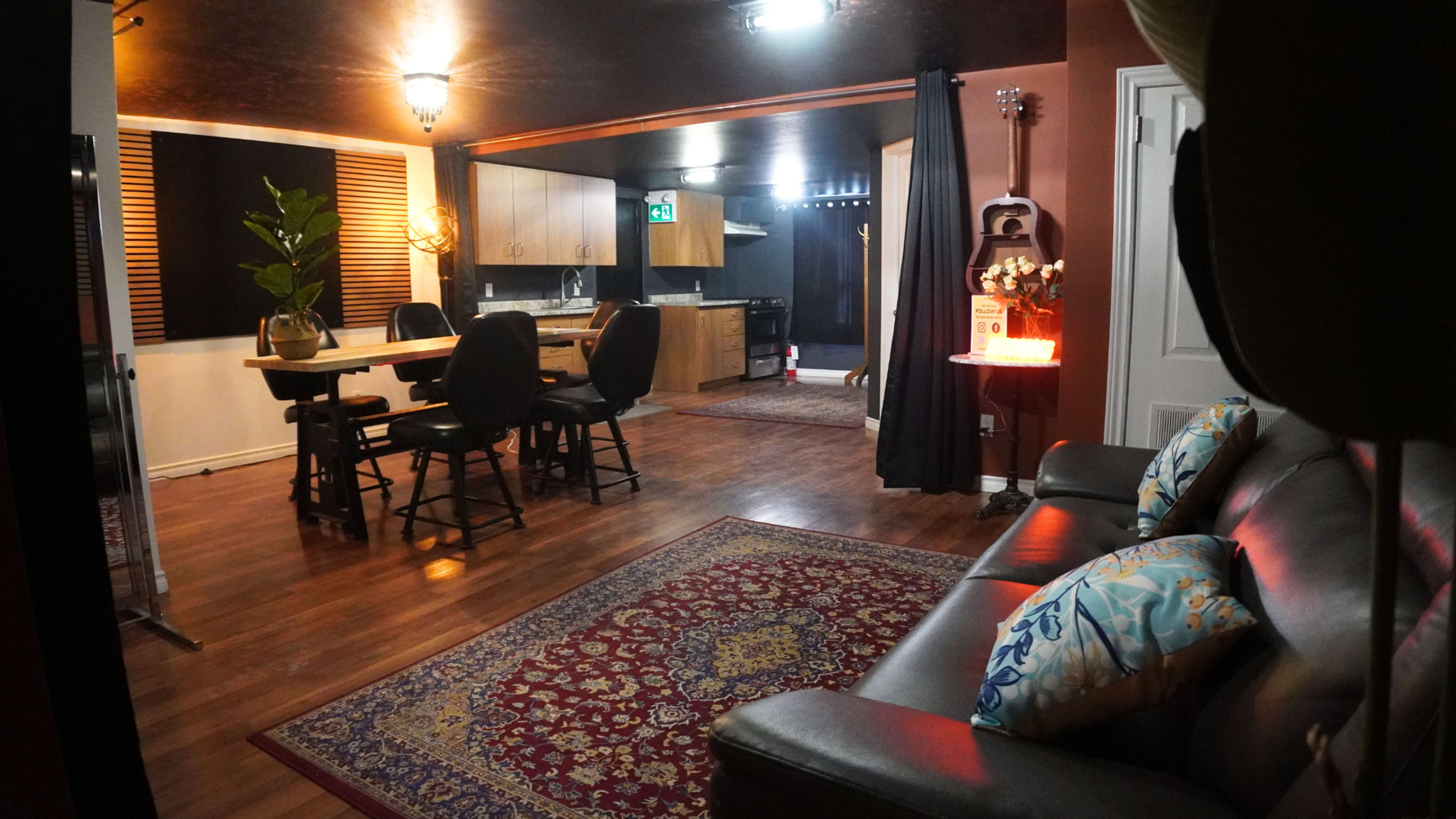 The image shows a cozy interior space with a living area featuring a dark sofa and decorative pillows, a dining table and chairs, and a kitchenette in the background.