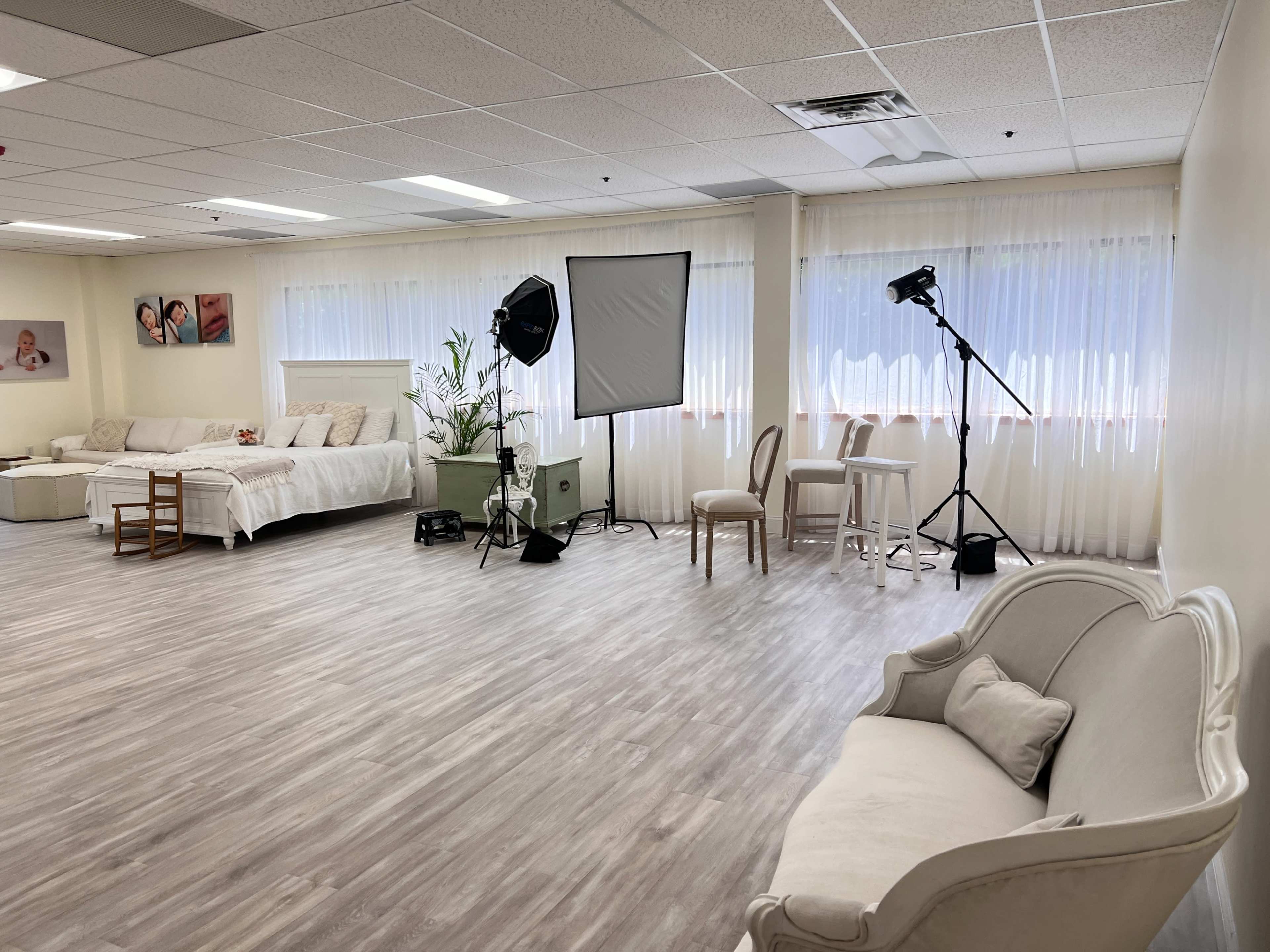 The image shows a spacious studio with a light-colored sofa, a bed adorned with white linens, photography equipment, and large windows with sheer curtains.