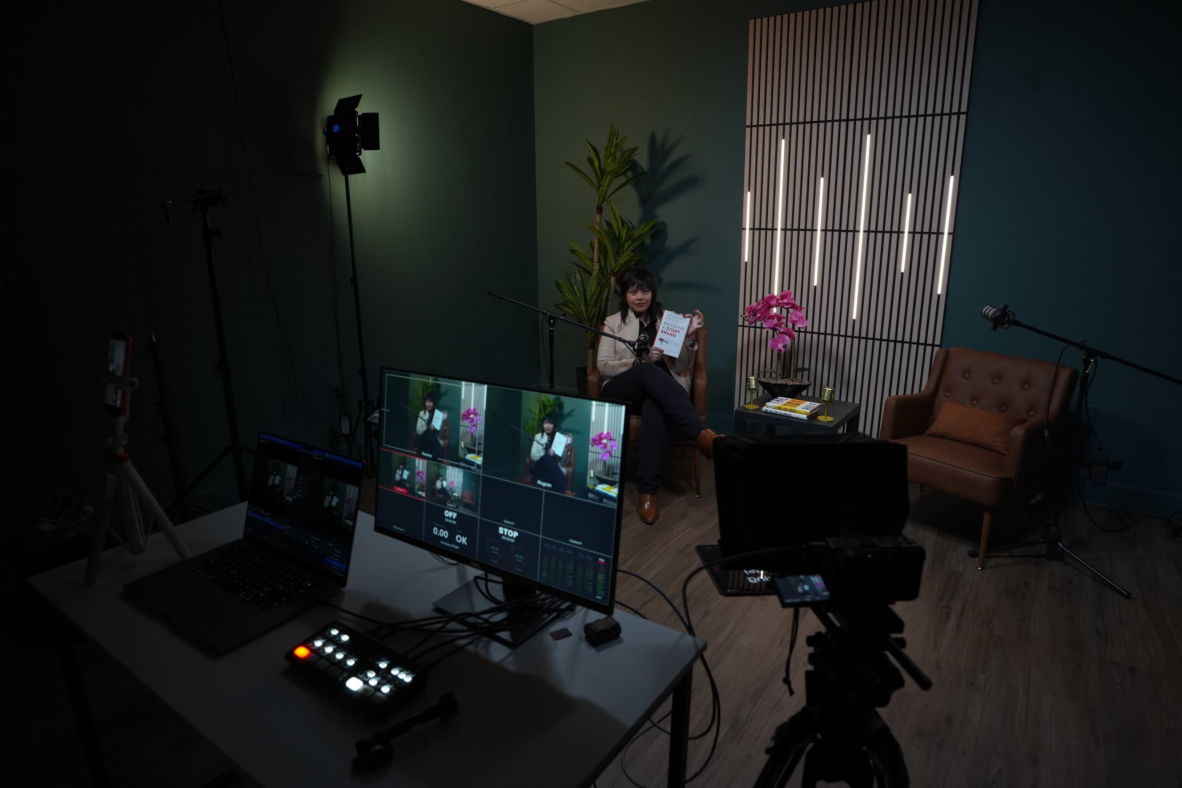 A person is seated in a studio setting equipped with cameras and lighting, holding a clipboard, while a plant and an orchid are visible in the background.