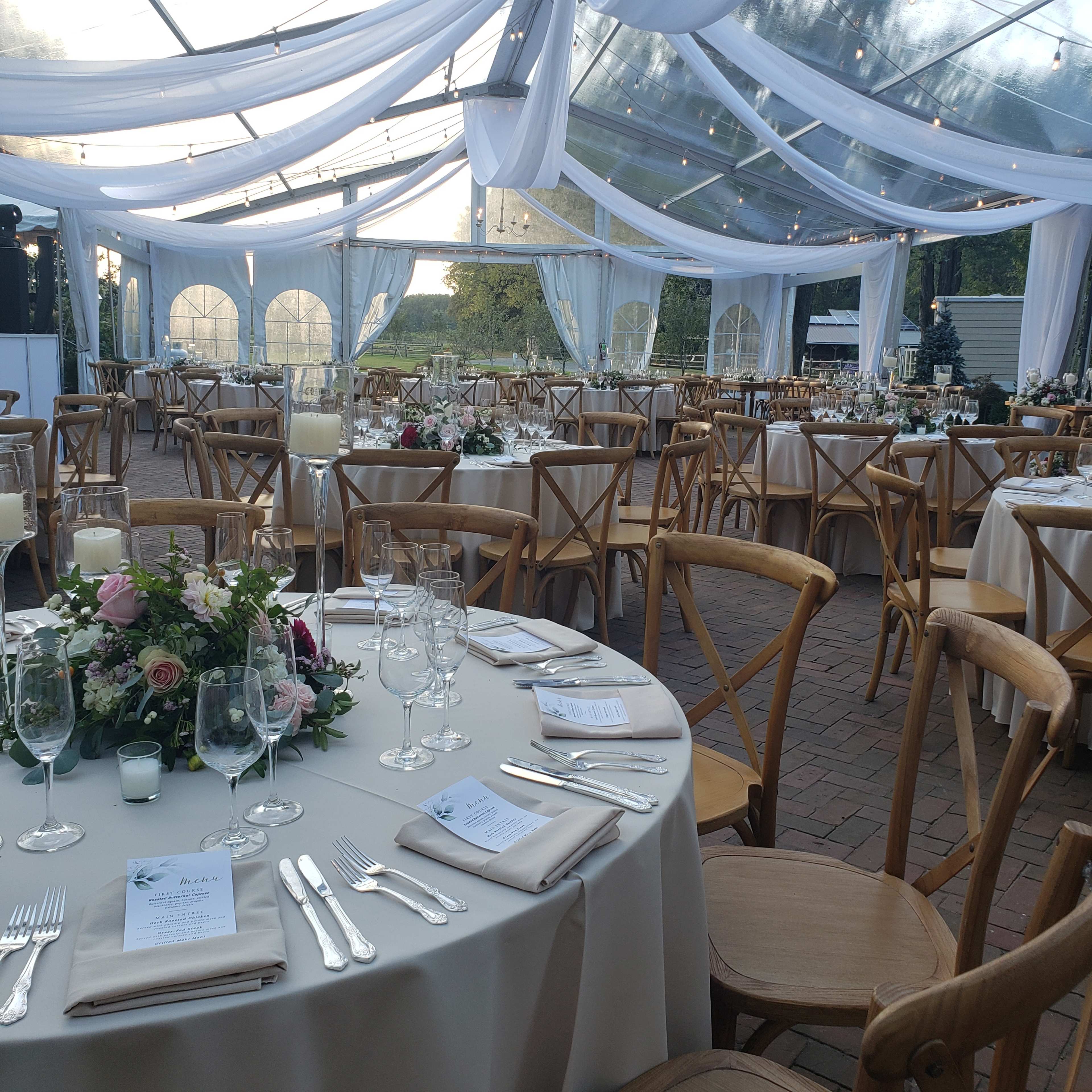 The scene shows a beautifully arranged wedding reception under a clear tent, featuring elegantly set tables adorned with floral centerpieces and glassware.