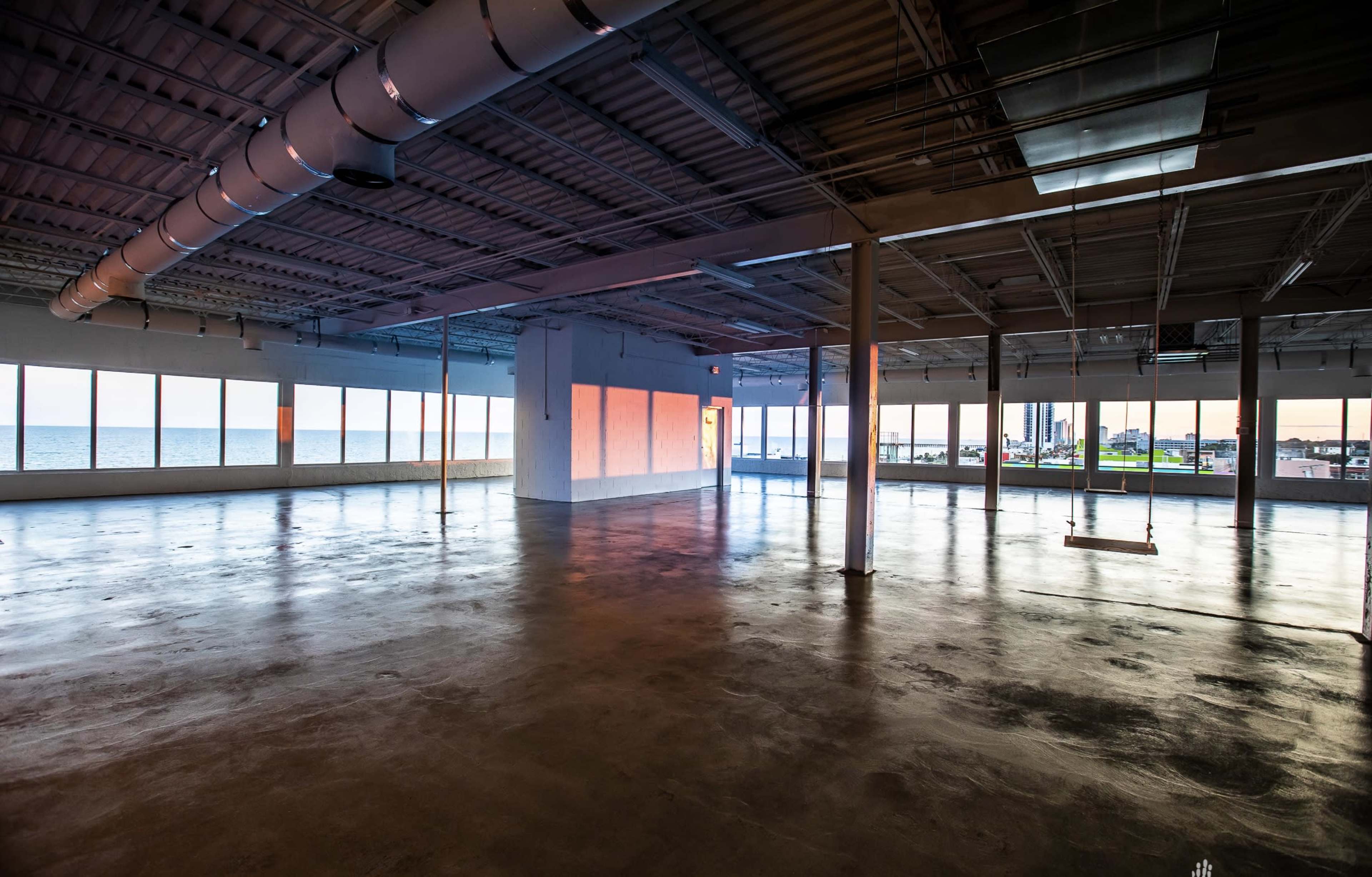 The image shows a spacious, empty room with large windows overlooking a body of water and a cityscape, illuminated by soft evening light.
