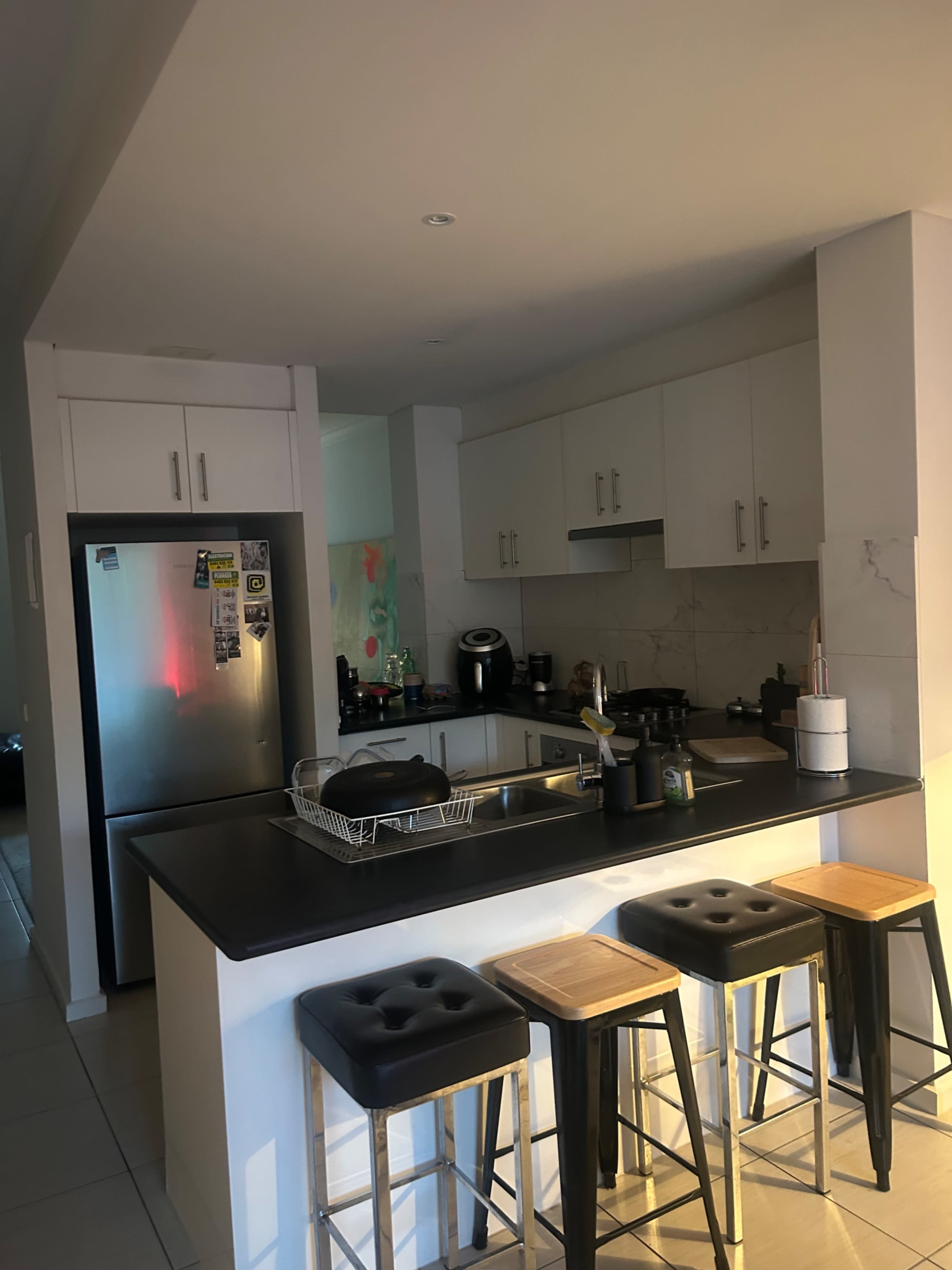 The image shows a modern kitchen with a marble countertop, stainless steel refrigerator, and four bar stools arranged in front of the island.
