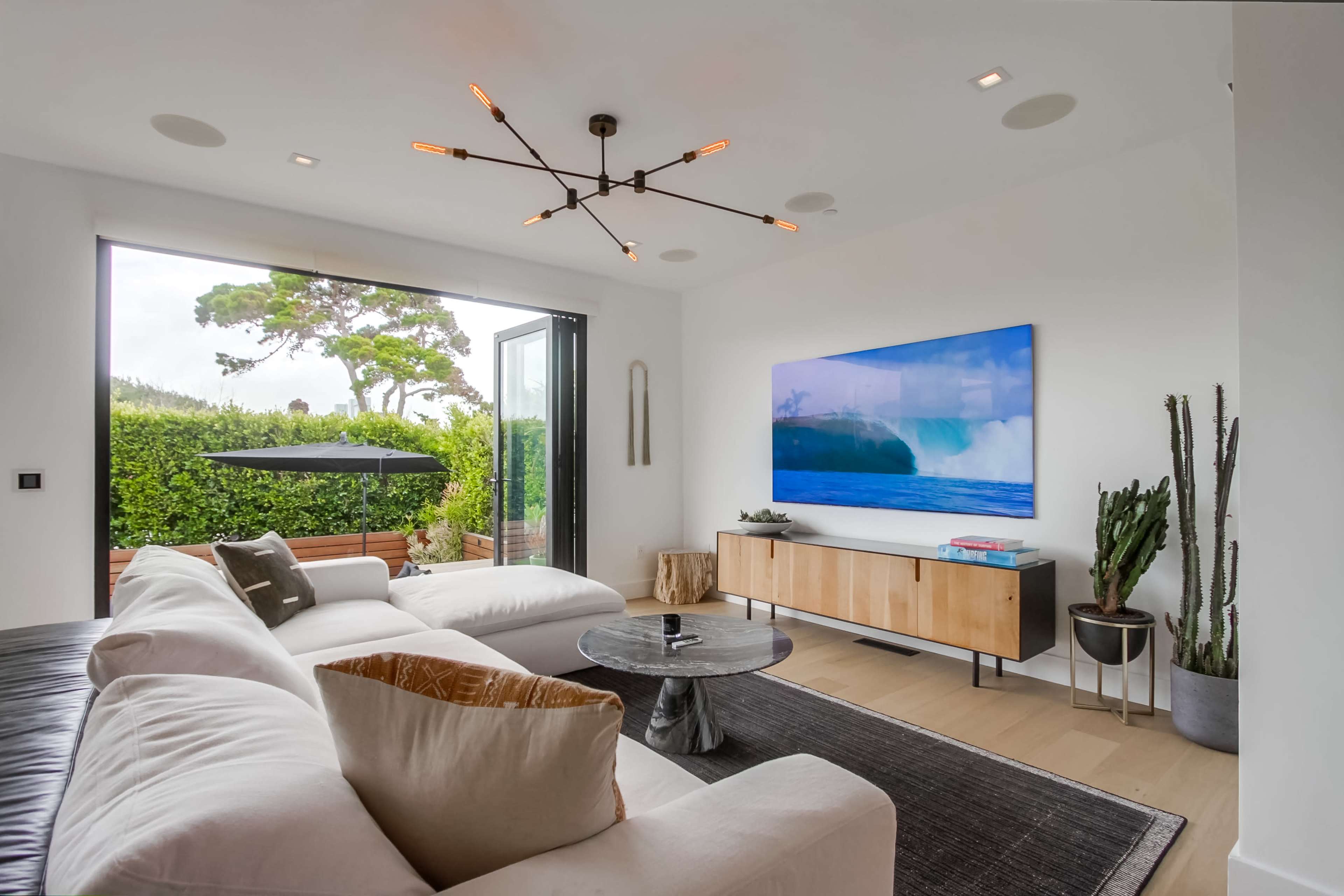 A modern living room with a large white sectional sofa, a round coffee table, a wall-mounted television displaying a scenic image, and large glass doors leading to an outdoor area.