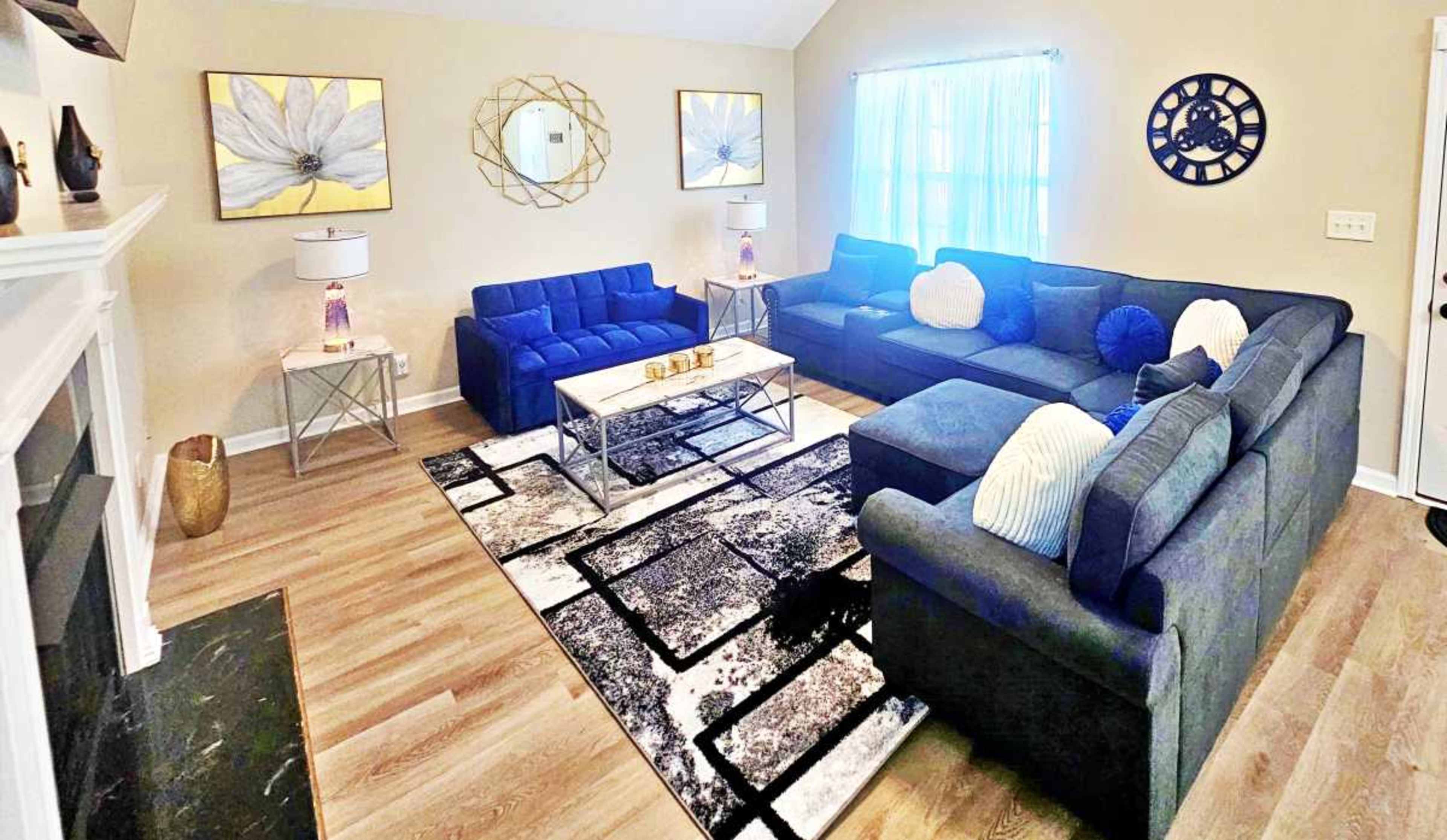 The living room features two blue sofas, a glass coffee table, and a patterned rug, with decorative wall art and soft lighting from a window.