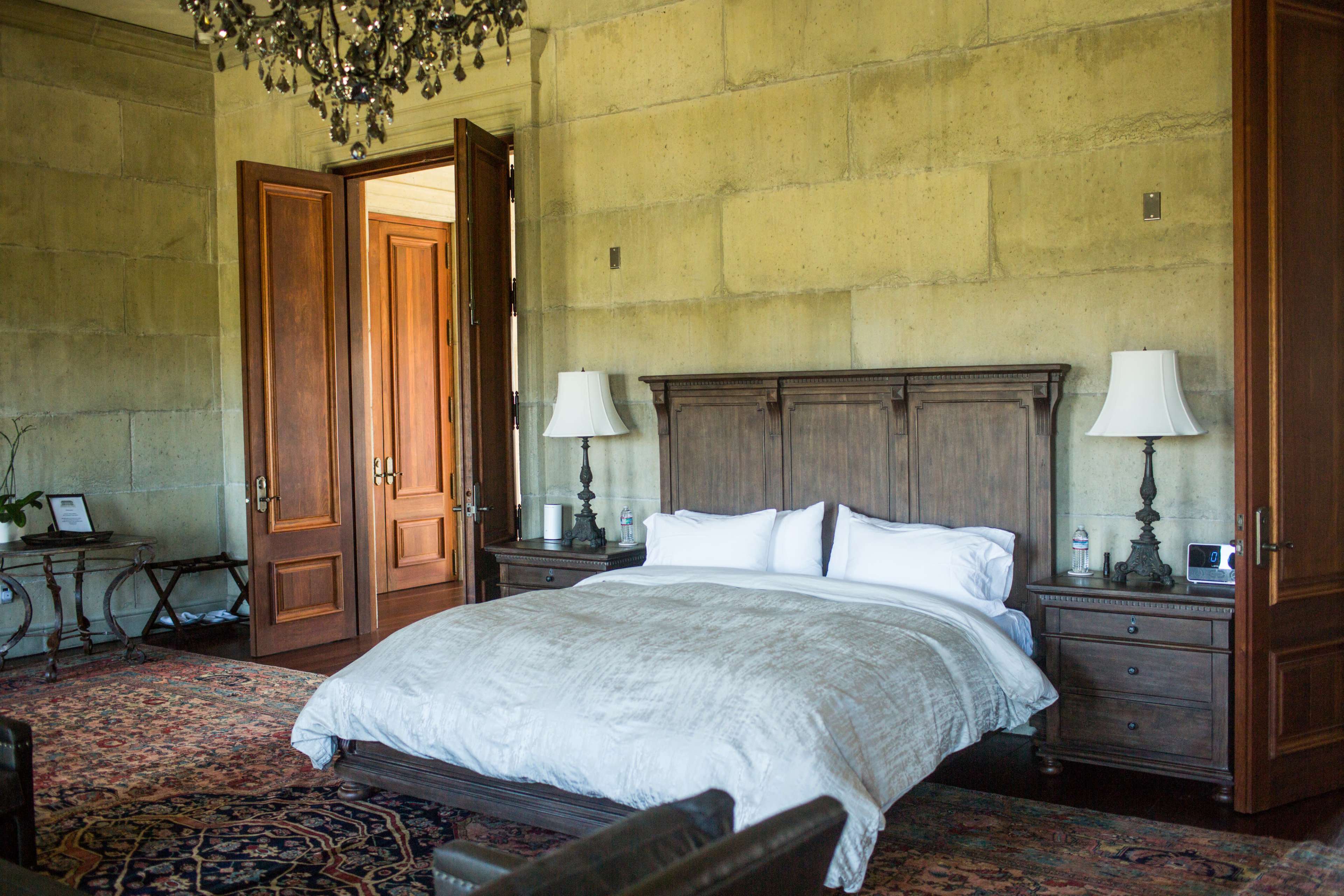 A spacious bedroom with a wooden bed, nightstands with lamps, and large wooden doors in a softly lit interior.