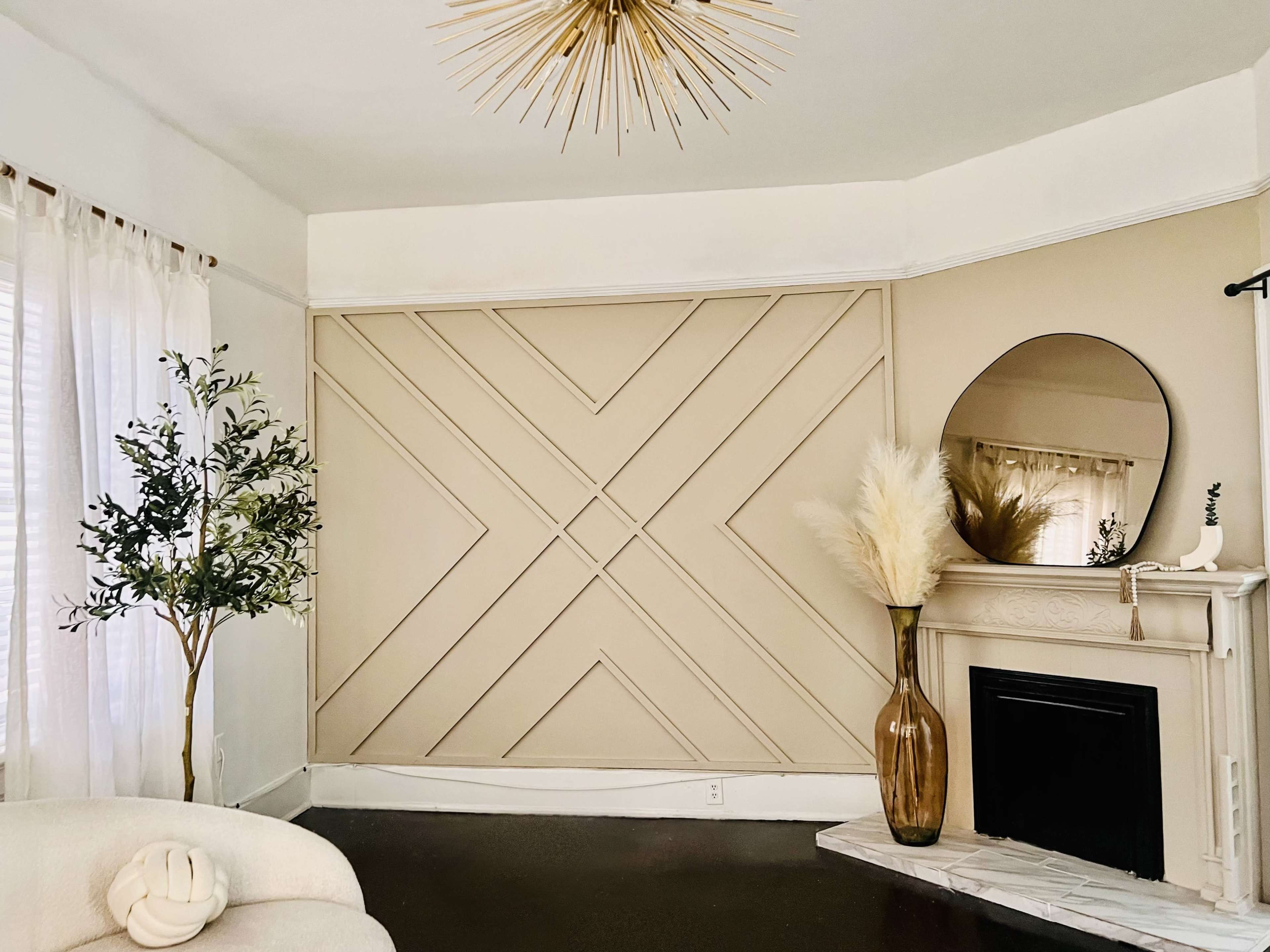 A modern living room features a neutral color scheme with a geometric wall design, a decorative mirror, a fireplace, and a tall vase containing pampas grass.
