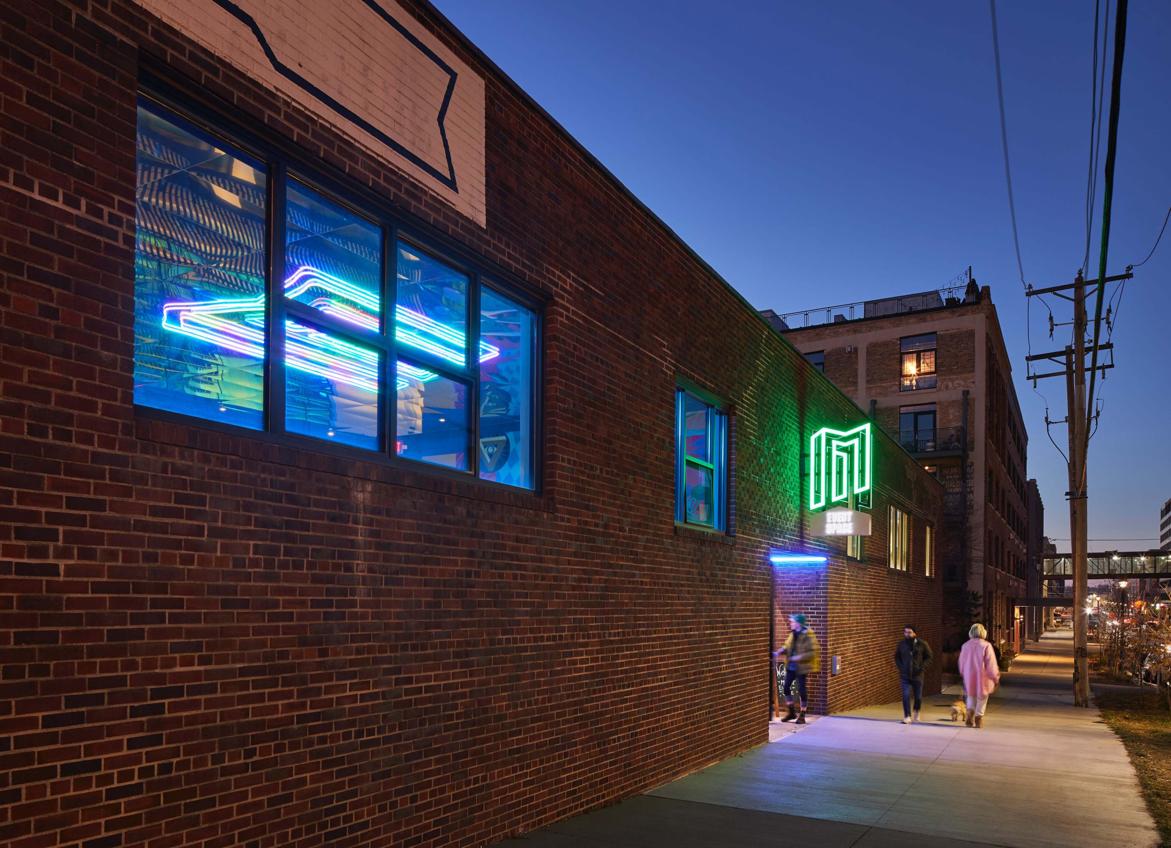 A brick building with illuminated neon signs is set against a twilight sky, while pedestrians walk along the sidewalk.