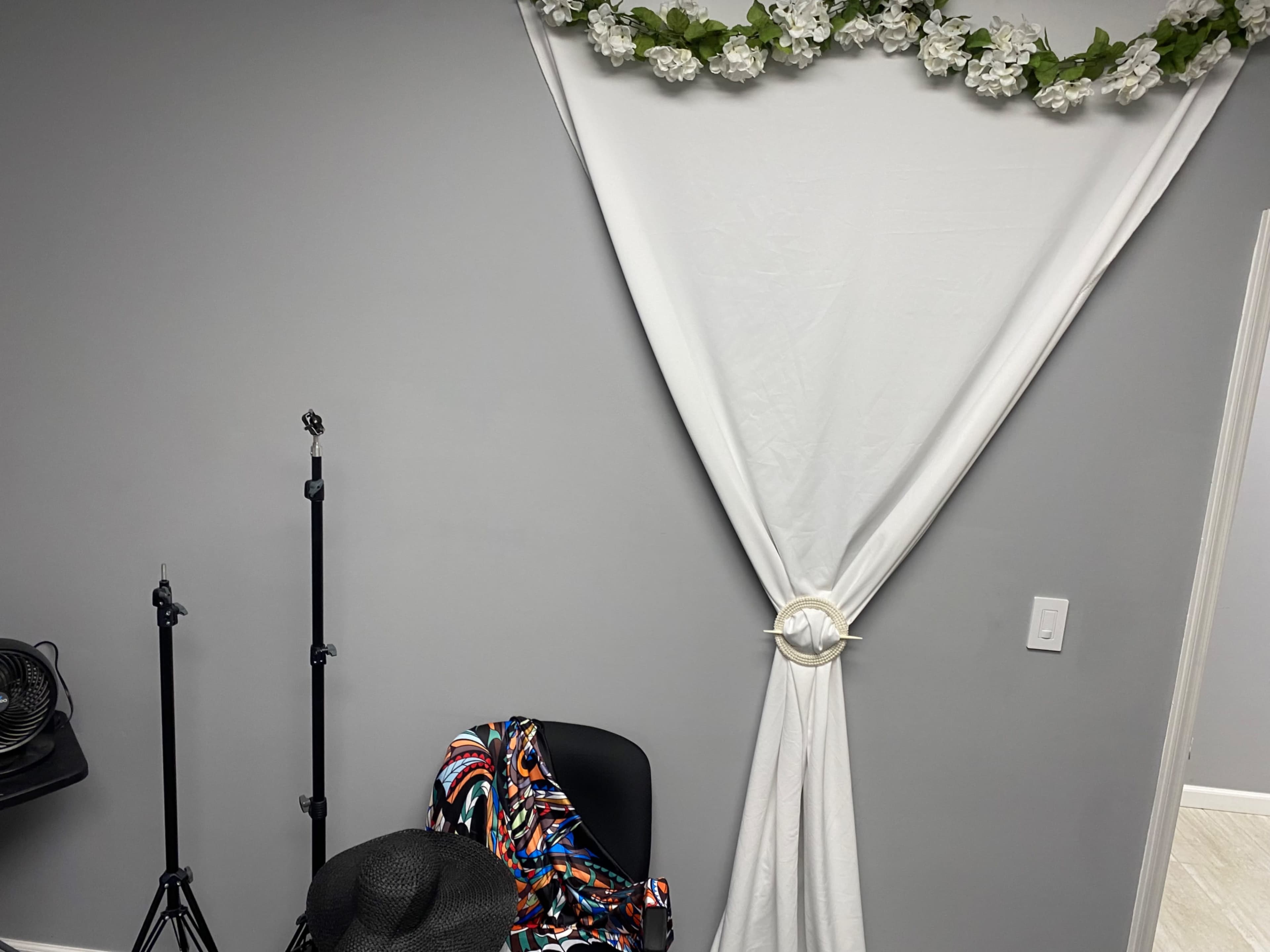 The image shows a gray wall adorned with a white drape and artificial flowers, next to a chair and a few stands.