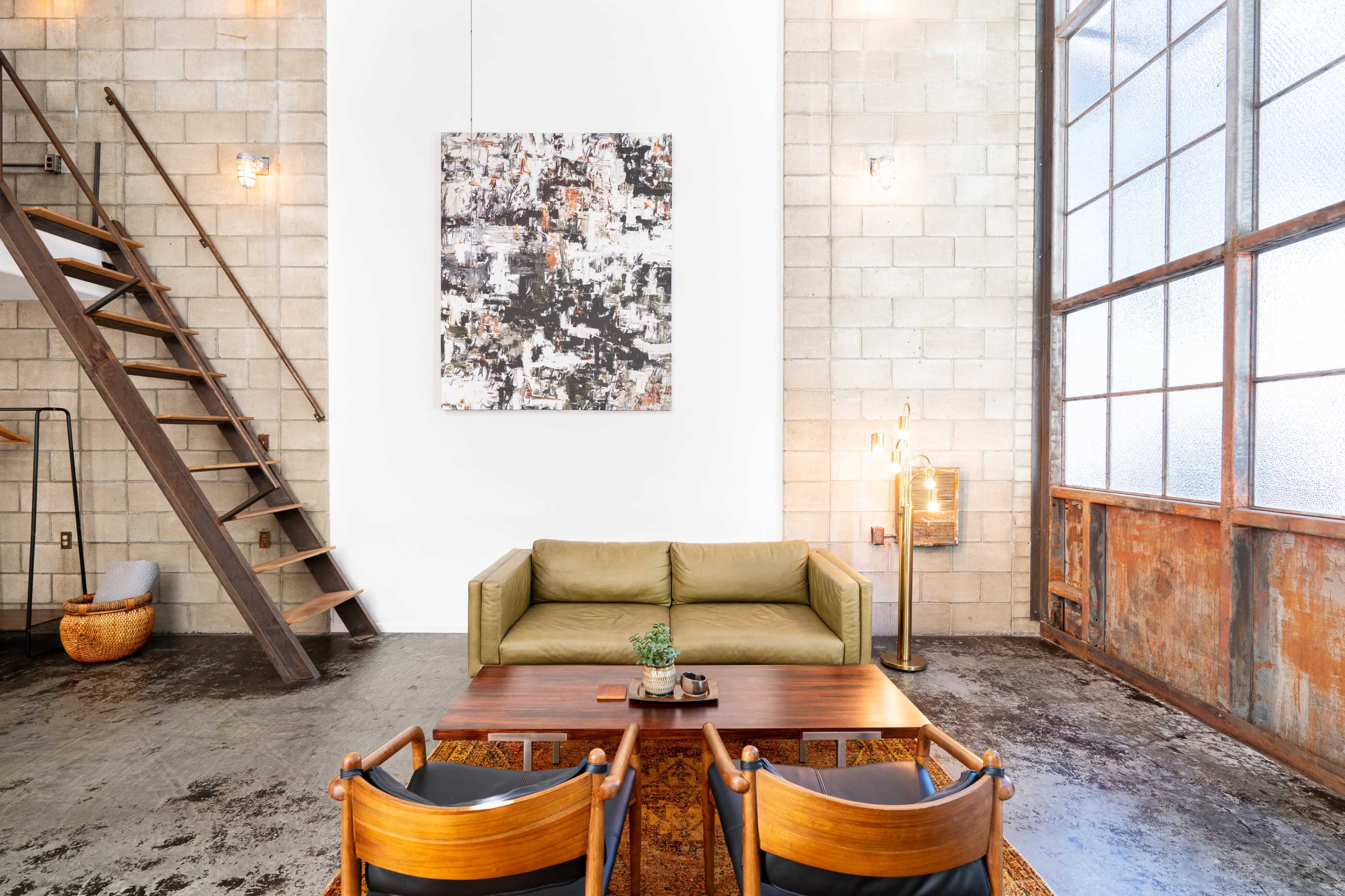 The image shows a modern living space with a green sofa, a wooden coffee table, and a large abstract painting on a white wall, alongside a staircase and large windows.