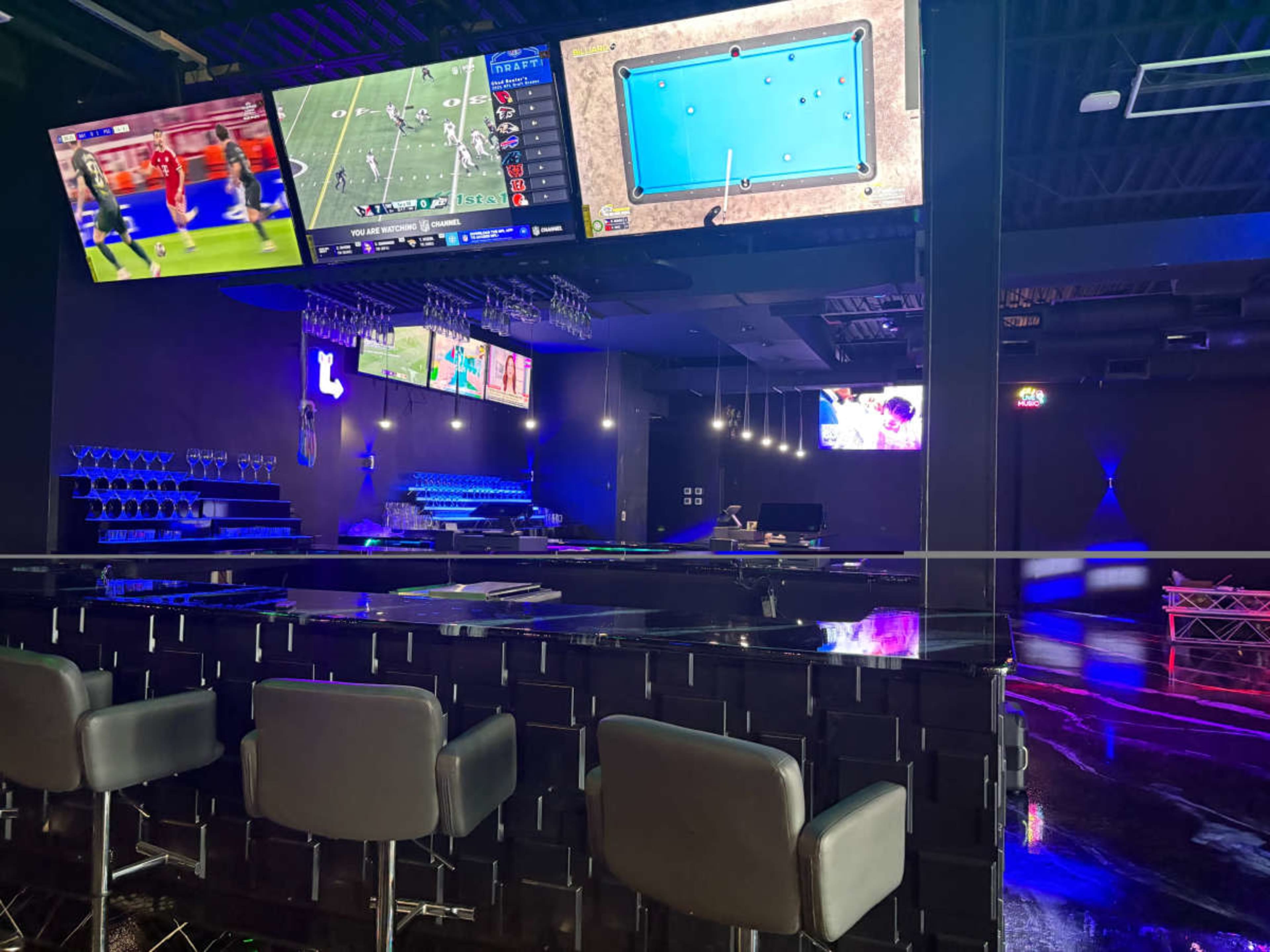 The image shows a modern bar area with multiple screens displaying sports events and a pool table, accompanied by sleek furnishings and dim lighting.